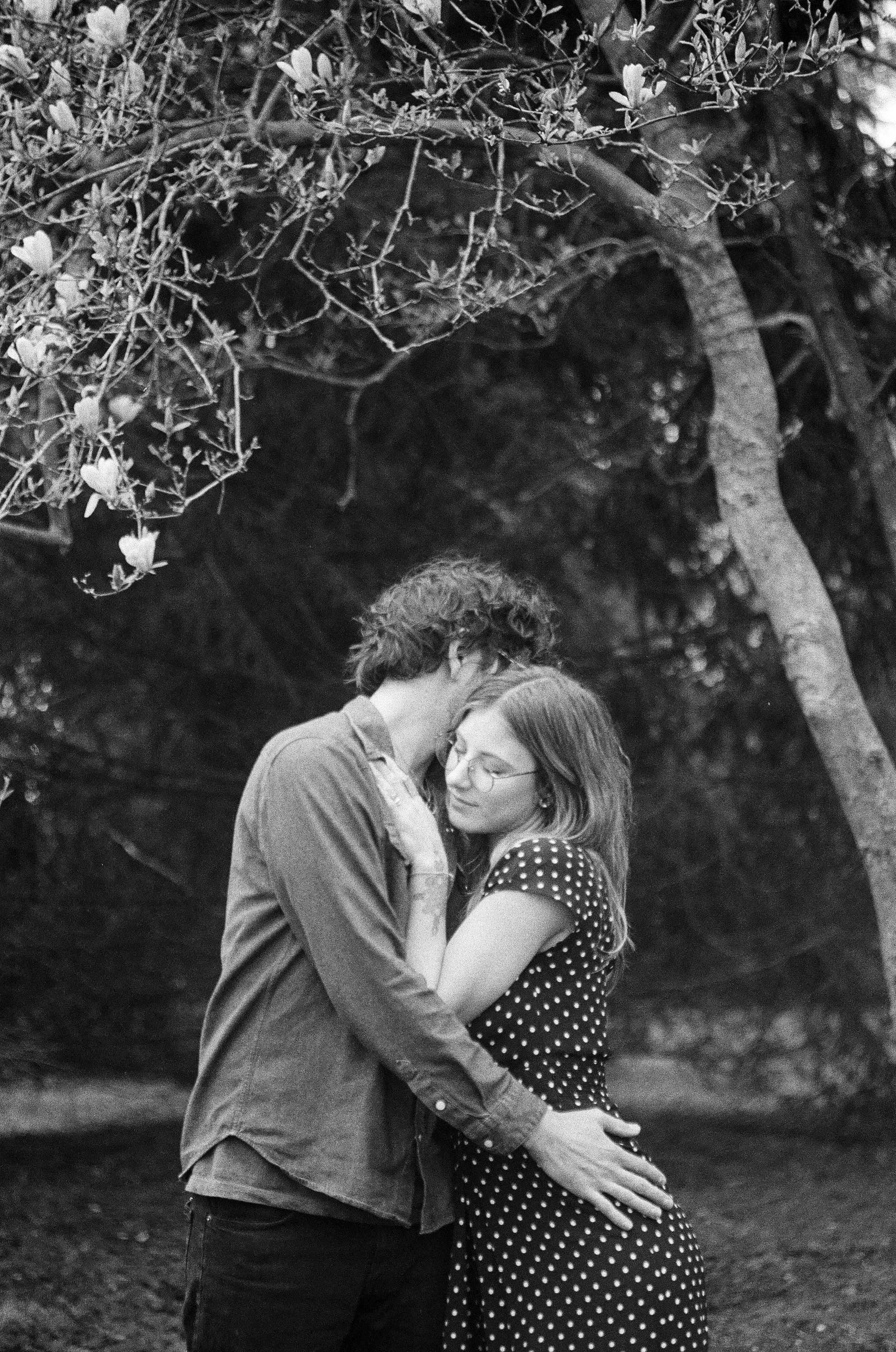 A black and white photo of a young couple embracing outdoors. The man and woman are holding each other closely, with the man's forehead touching the woman's forehead and their eyes closed. They are surrounded by trees and natural foliage.