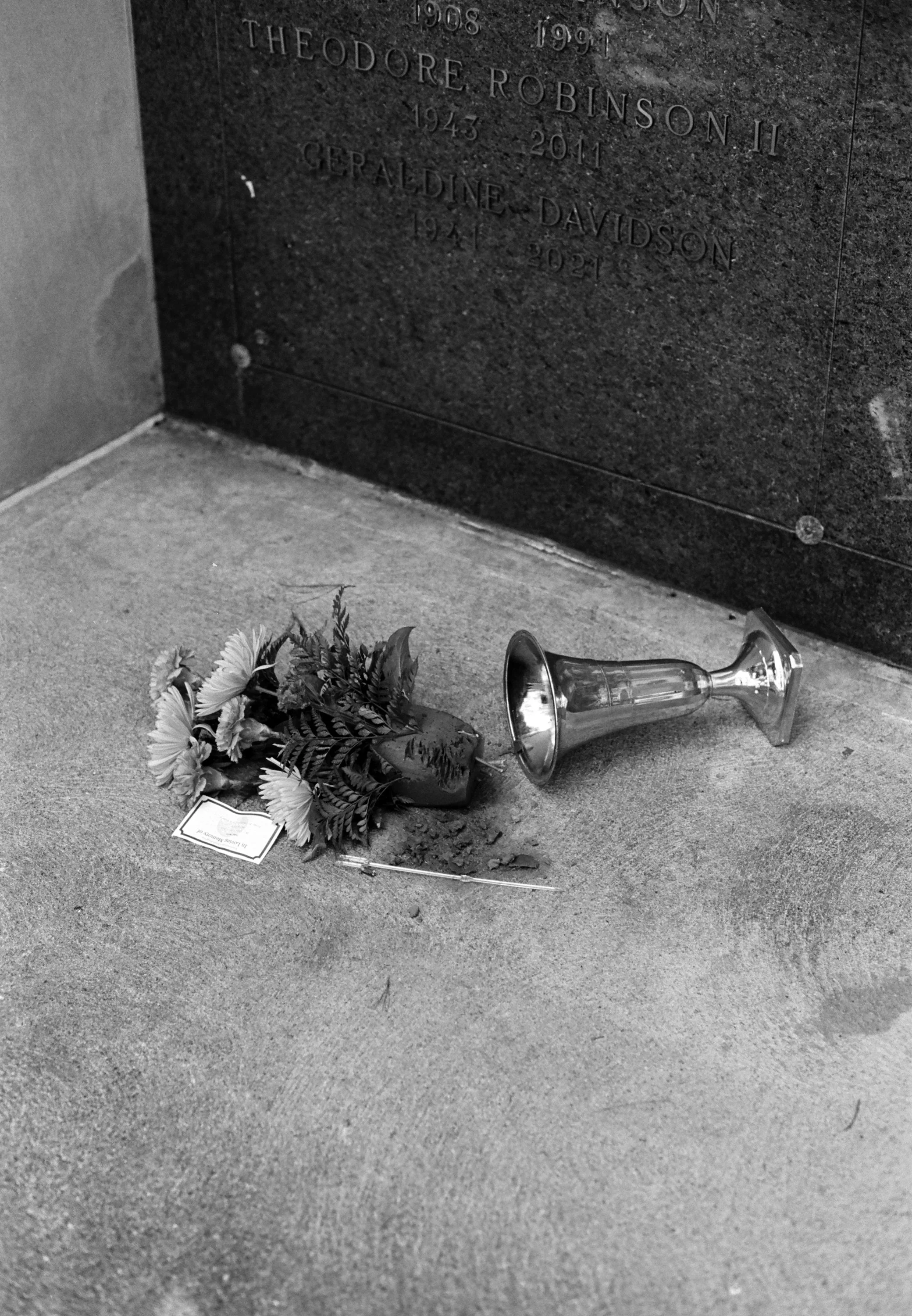 A memorial plaque on a black stone wall with engraved names and dates. On the ground, there is a bouquet of flowers in a container, a small card, and a silver trophy laying on its side.