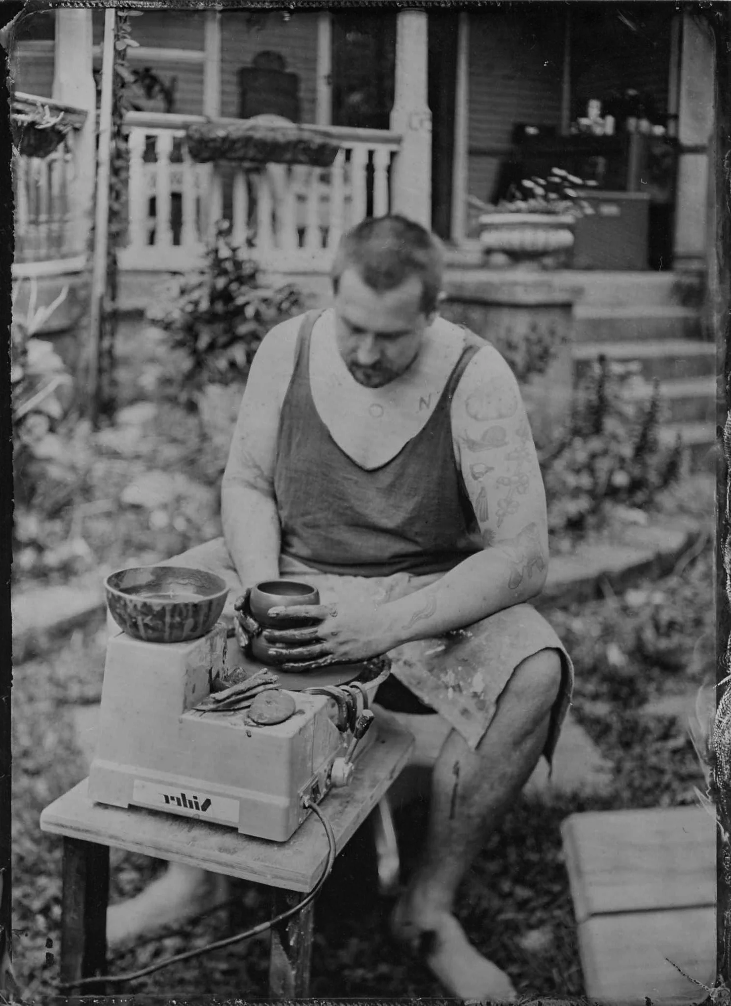 Black and white photo of a man sitting at a table outdoors, using a portable stove to cook. He has tattoos on his left arm, wears a tank top, and is holding a cup. The background shows a porch with a railing, steps, and a small house with shelves and
