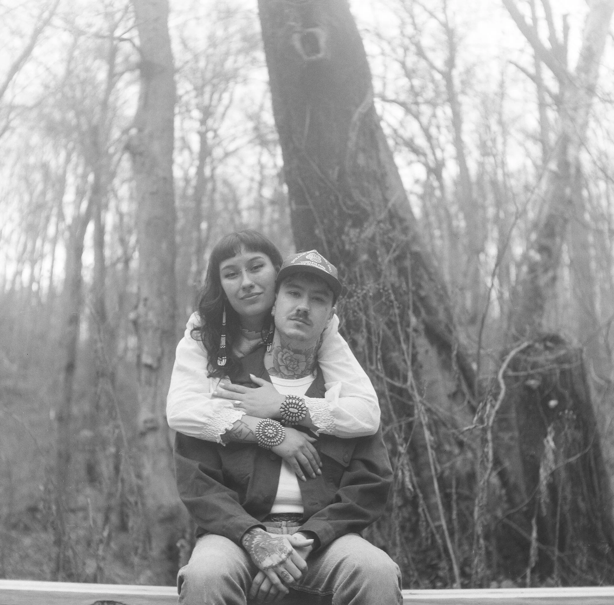 Two children sitting on the ground in a forest with trees in the background, one girl with dark hair and a boy wearing a cap.
