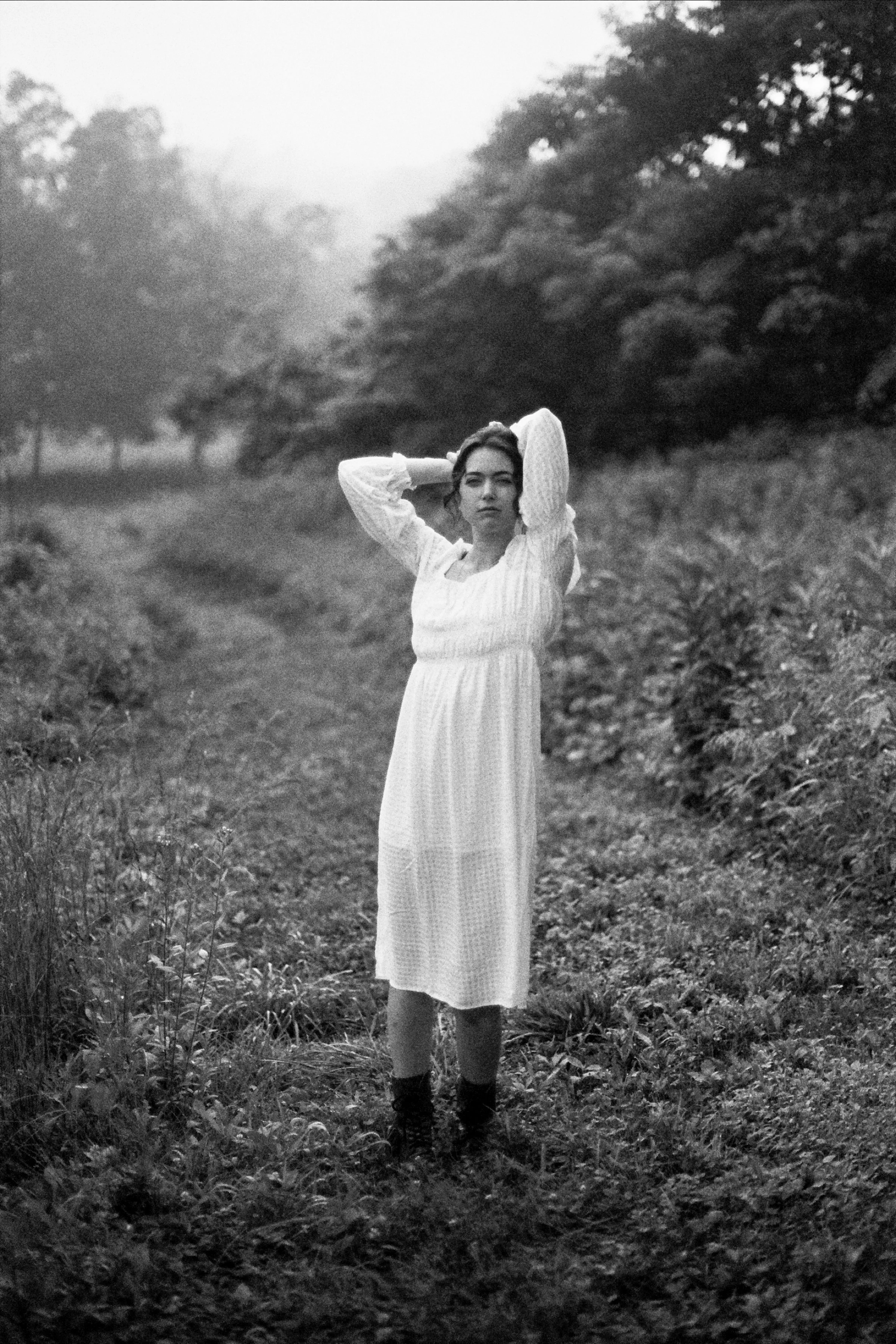 A woman in a long, white dress standing in a natural outdoor setting with trees and grass, looking directly at the camera, with her hands behind her head.