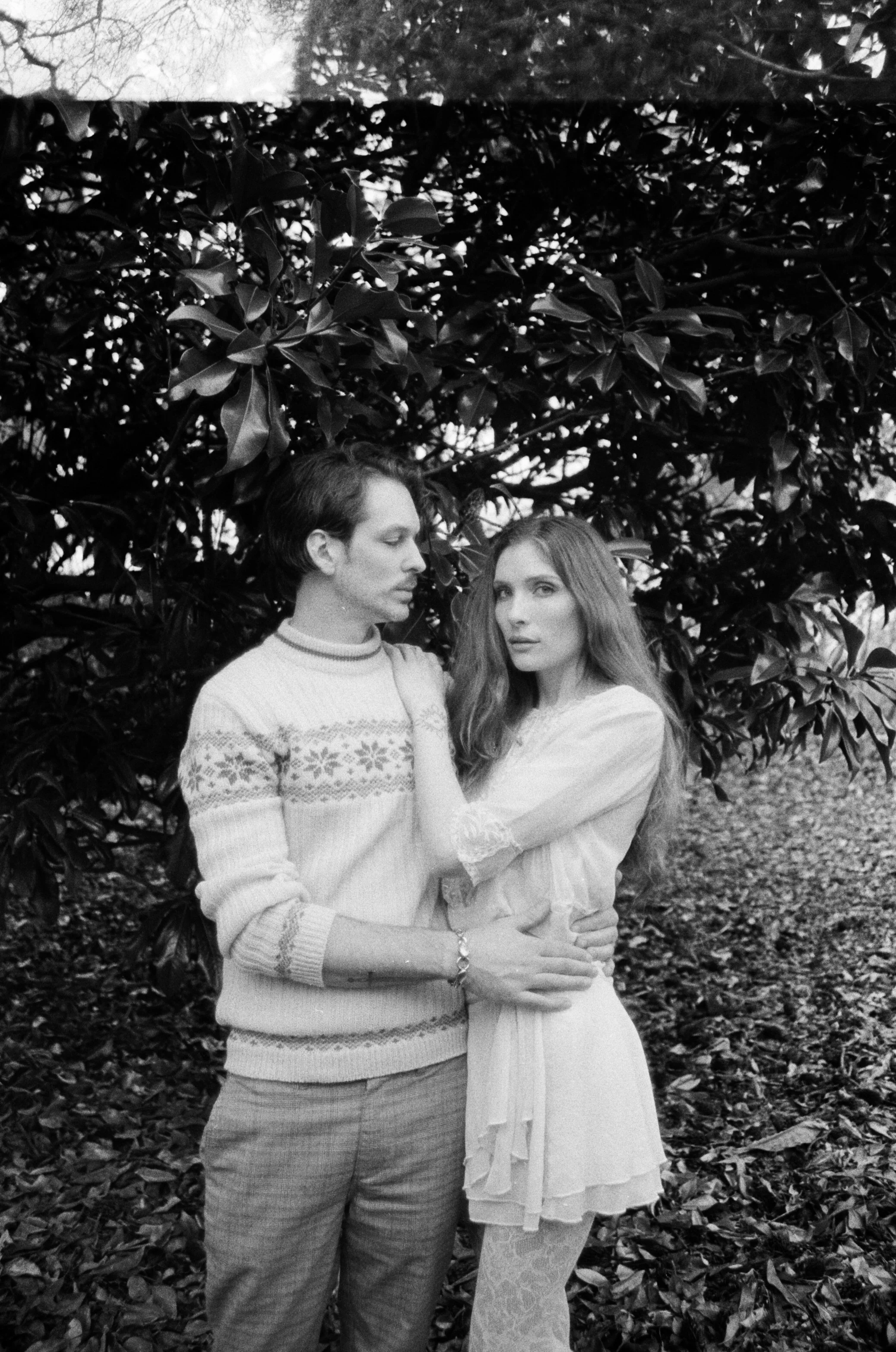 A black and white photo of a man and woman standing outdoors among trees and fallen leaves. The man has dark hair, wears a patterned sweater and plaid pants, and is looking at the woman. The woman has long hair, wear a white dress with lace details, 