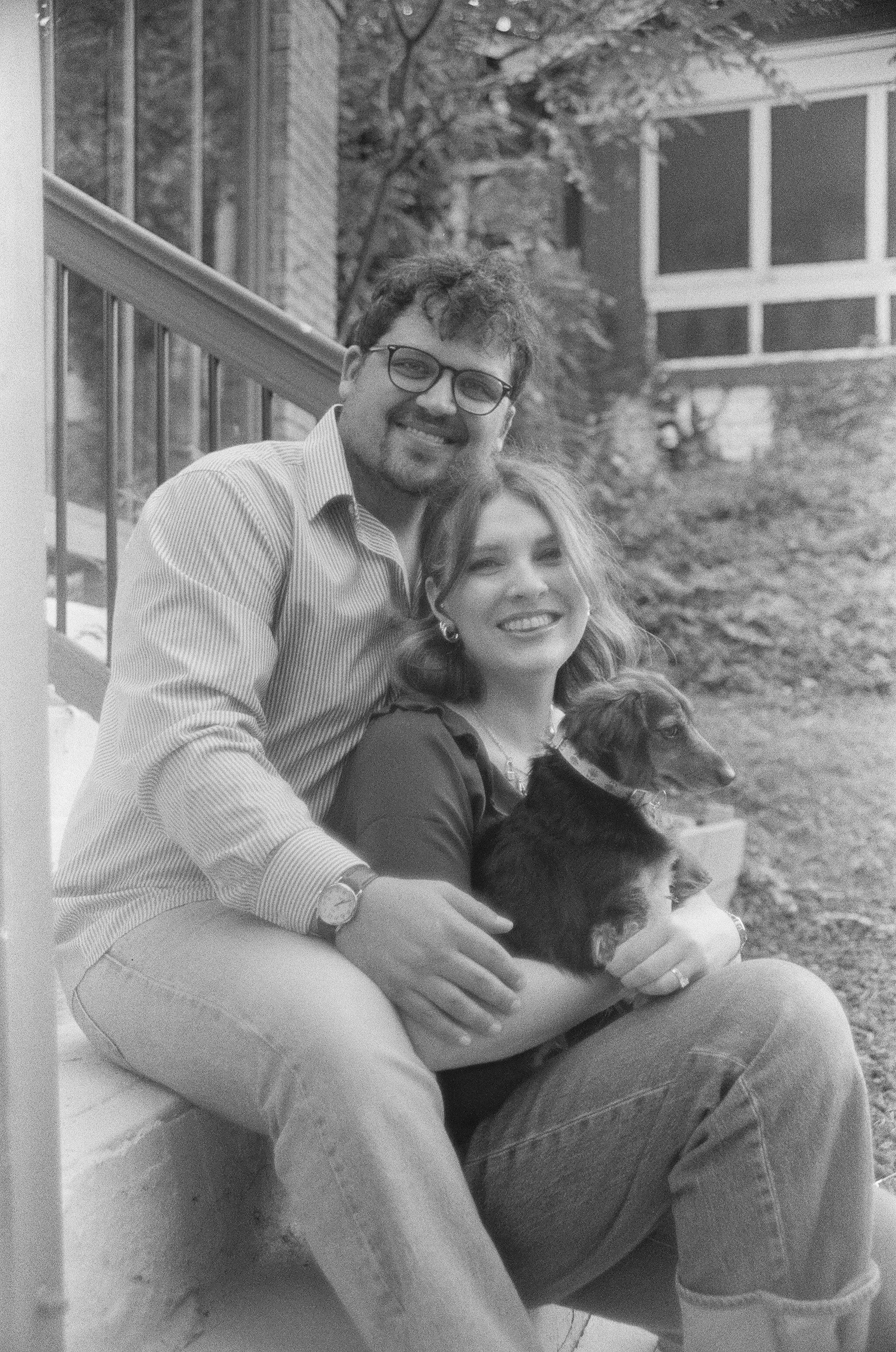 A black and white photo of a man and a woman sitting outside on a bench, holding a dog. The man has curly hair, glasses, a striped shirt, and is smiling. The woman has wavy hair, earrings, and is also smiling. They are near a wooden building with win