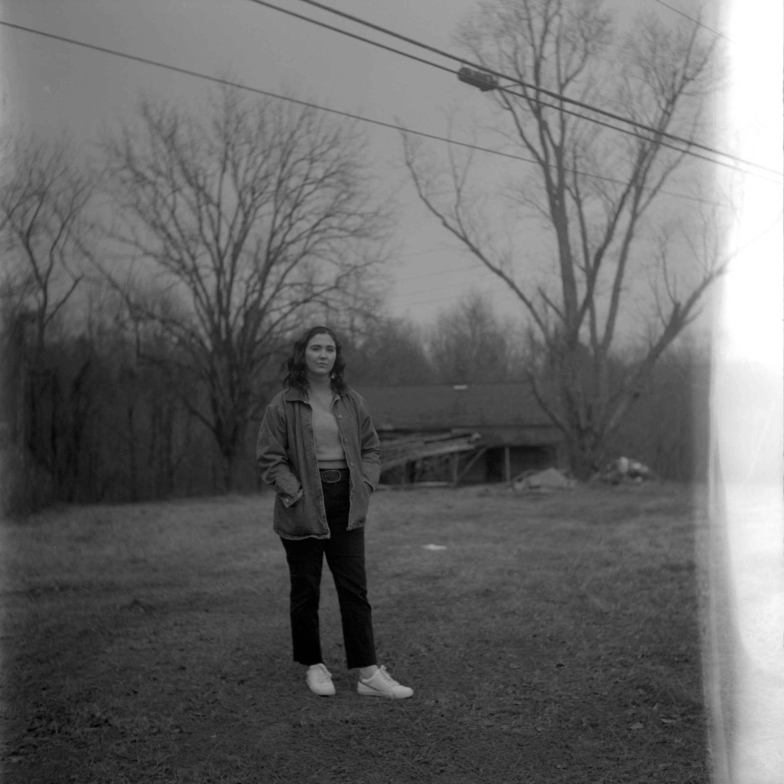 Black and white photo of a young woman with wavy hair standing outdoors on grass with leafless trees and power lines in the background. She is wearing a jacket, pants, and sneakers.