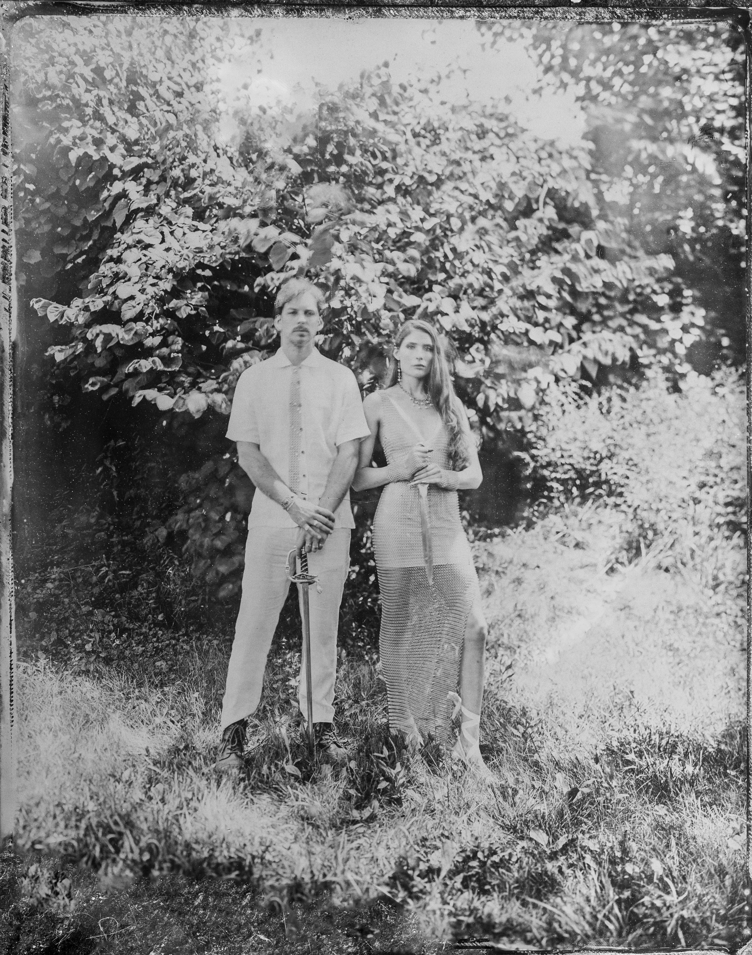 A black and white photo of a man and woman standing outdoors in front of leafy trees, with grass and plants on the ground.
