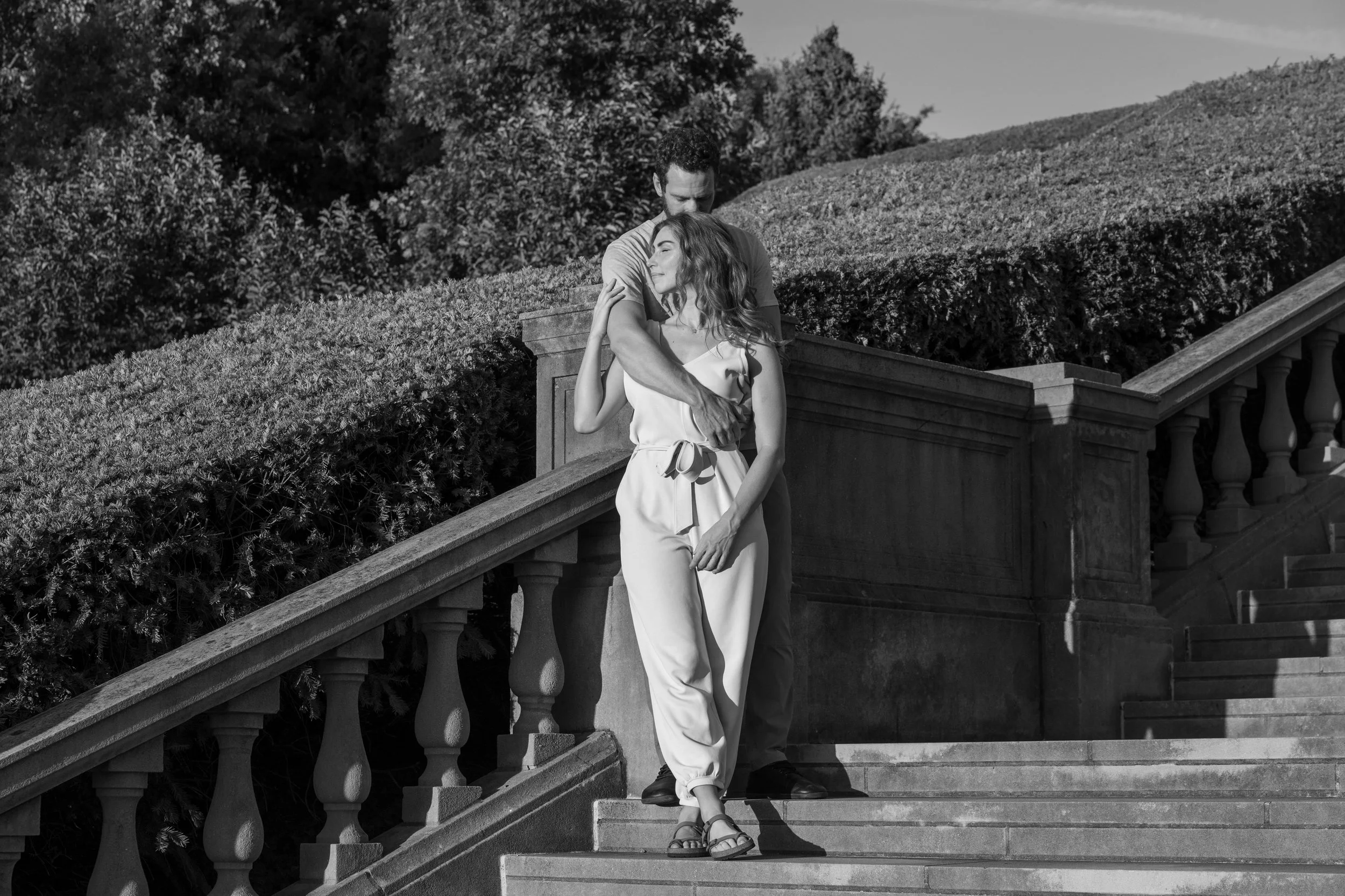 A black and white photo of a man and woman standing on outdoor stairs; the man is embracing the woman from behind while she rests her head on his chest, with trees and bushes in the background.