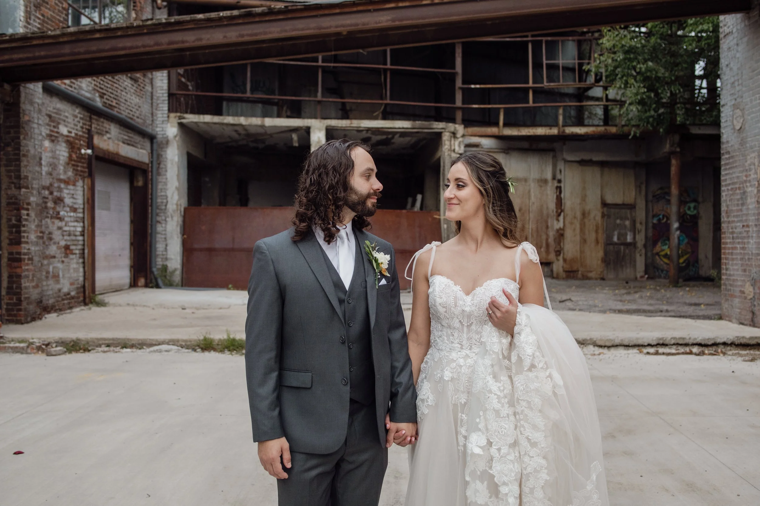 A bride and groom holding hands and looking at each other in front of an old urban building with exposed brick and metal framework.