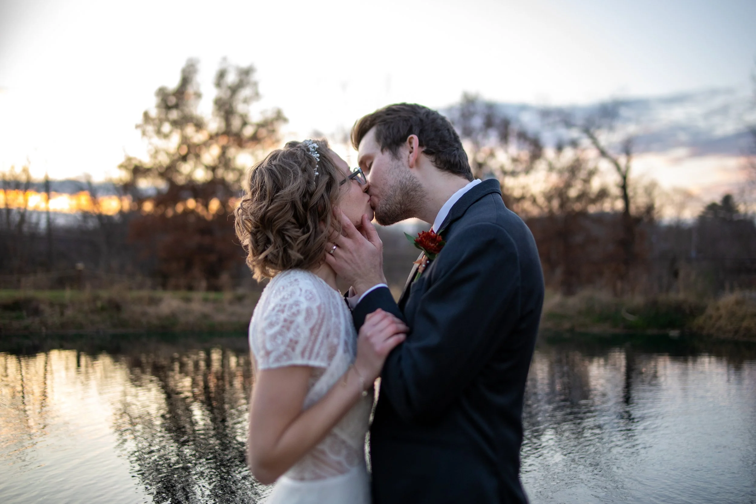 A couple sharing a kiss by a body of water during sunset, with trees in the background.