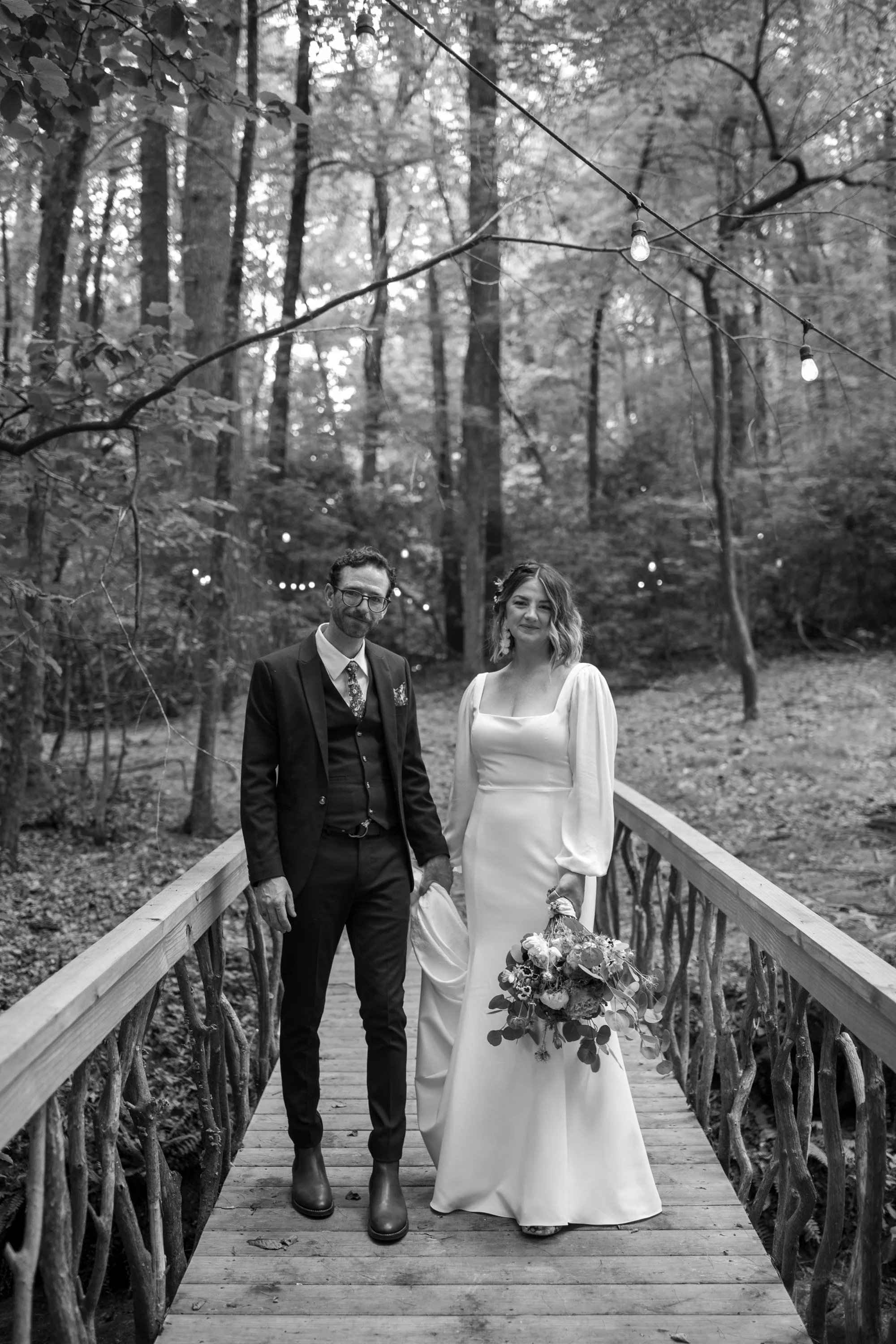 A couple dressed in wedding attire standing on a wooden bridge in a forested area with hanging string lights in the background.