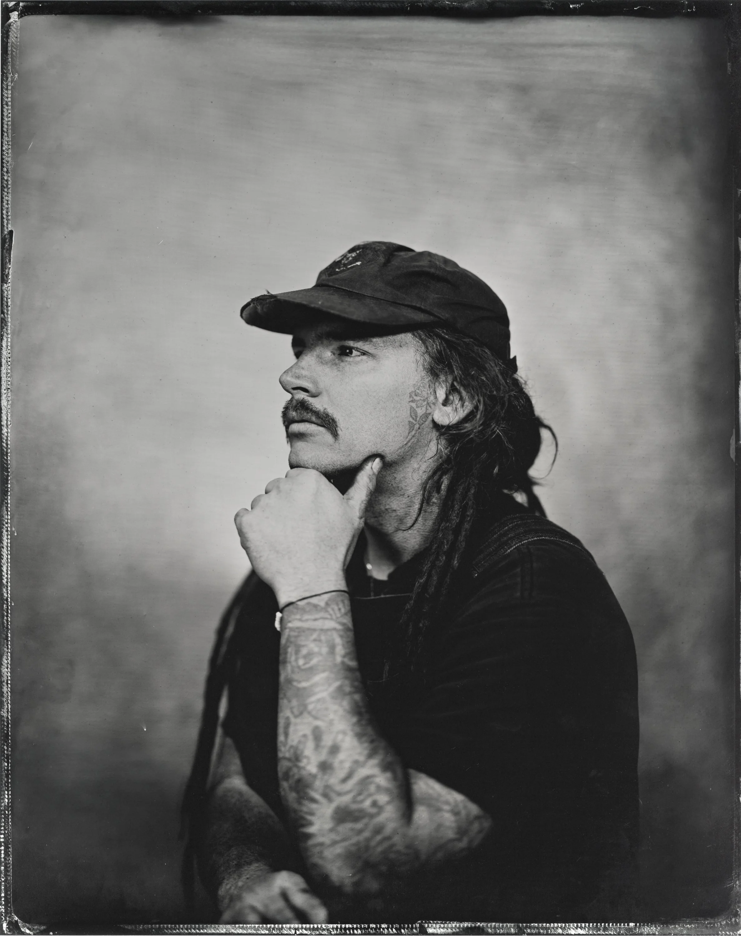 Black and white photograph of a man with long hair, a mustache, and beard wearing a baseball cap. He's resting his chin on his hand, with his fingers touching his cheek, and has tattoos visible on his arm and neck. The background is plain and slightl