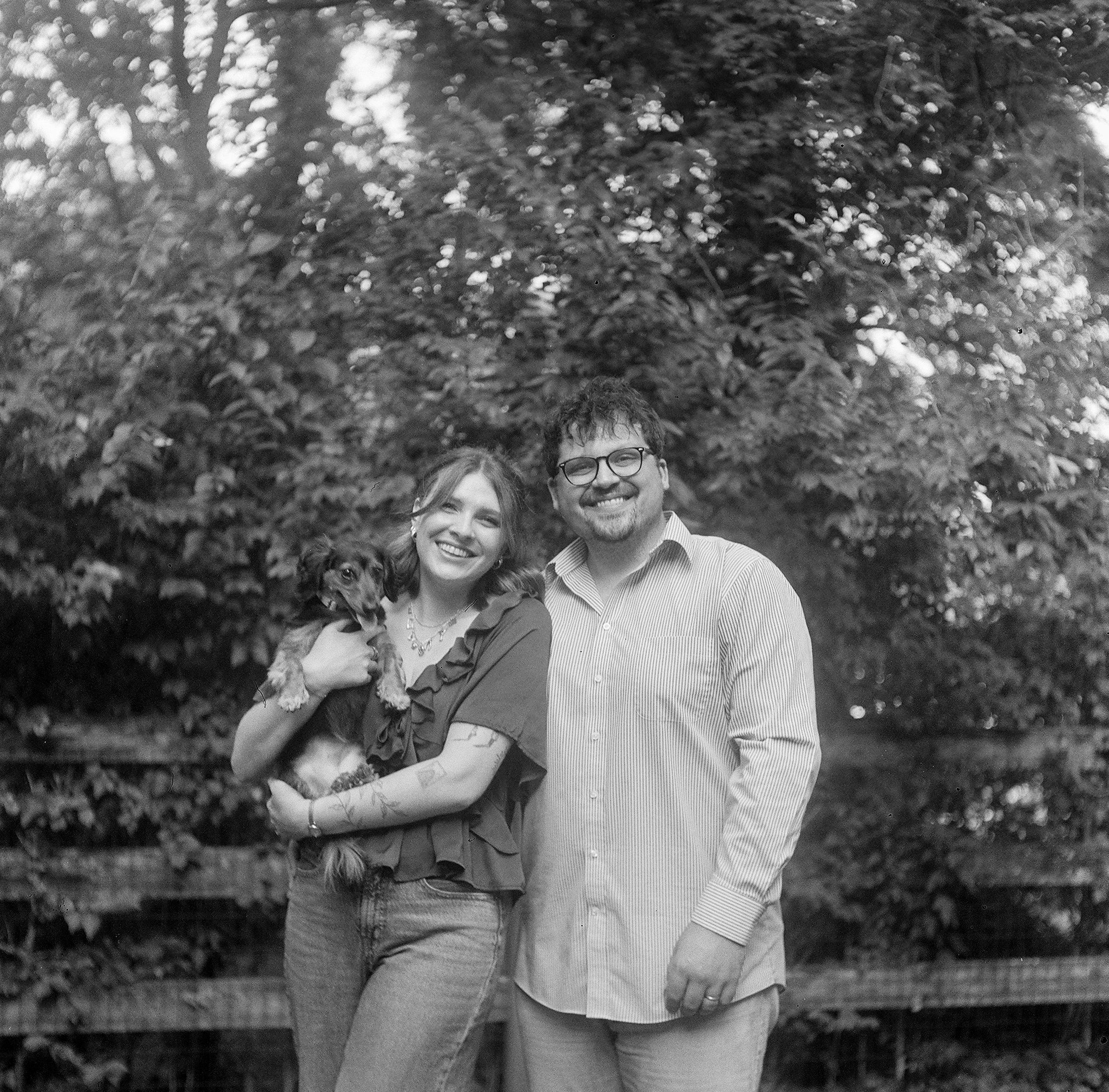 A smiling couple standing outdoors among trees, with a dog in the woman's arms, all appearing happy and close together.