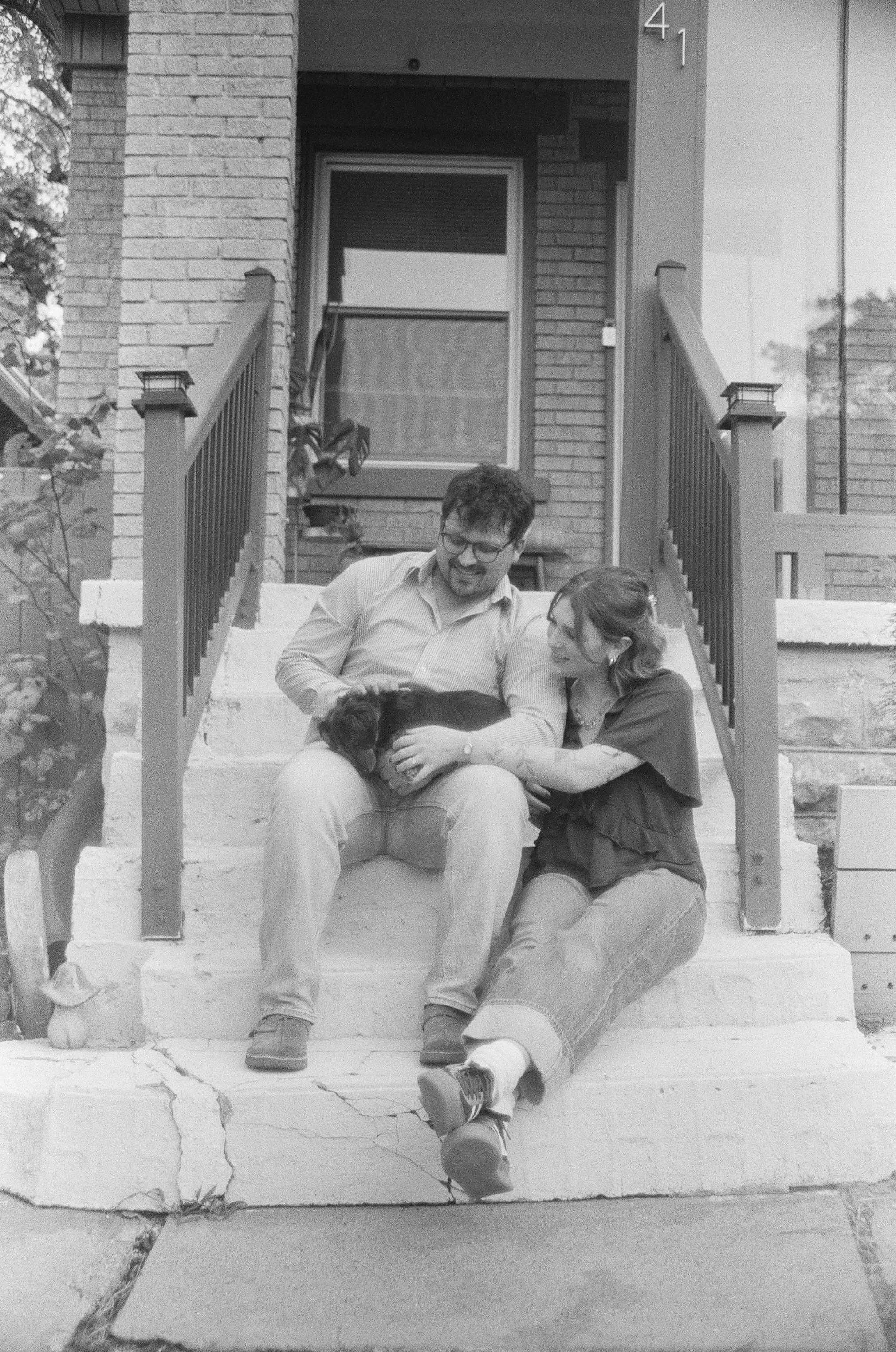 A man and a woman sitting on steps in front of a brick house, holding a small puppy. The man is wearing glasses and a striped shirt. The woman is leaning toward the man, smiling. The house has the number 41 on it, and there are plants next to the ste