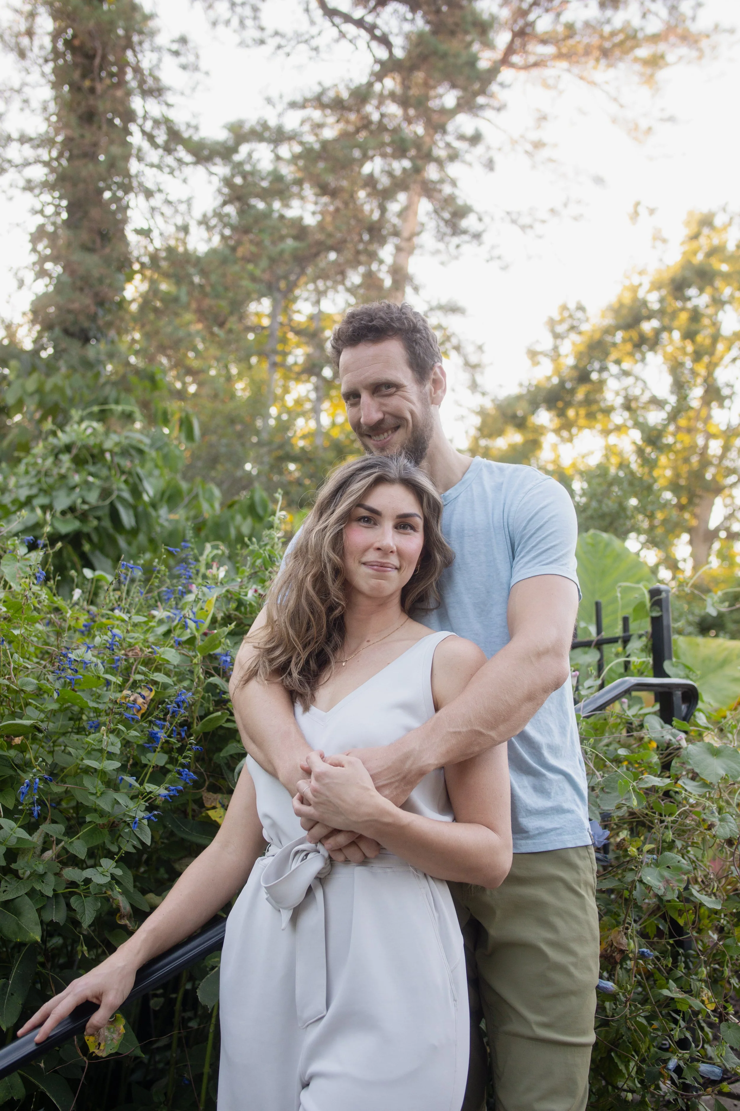 A man and woman embracing each other outdoors in a garden, with trees and foliage in the background, during daylight.