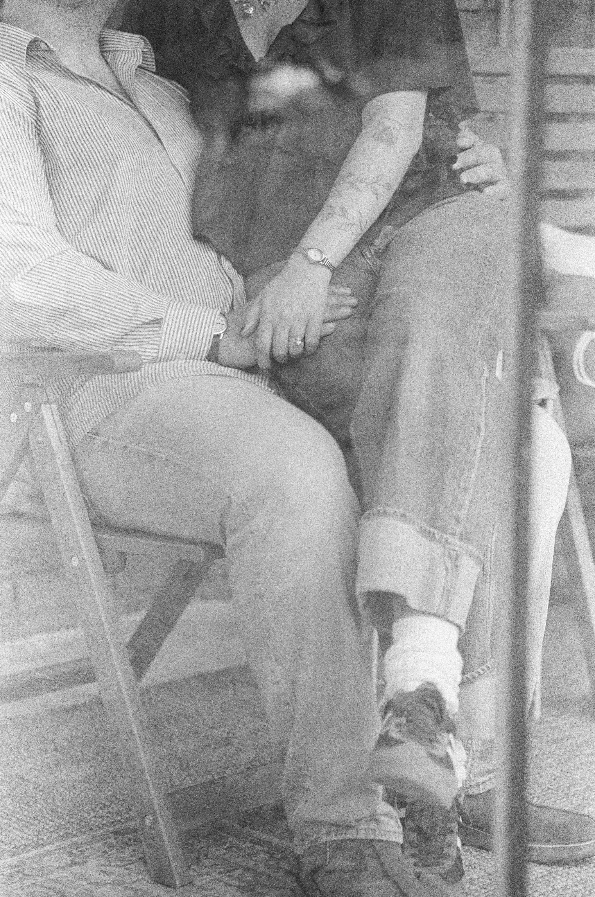 Two people sitting closely together on chairs, with one person's arm around the other. The person on the left is wearing a striped shirt and sitting on a wooden chair, while the person on the right is wearing jeans and sneakers, sitting on a chair wi