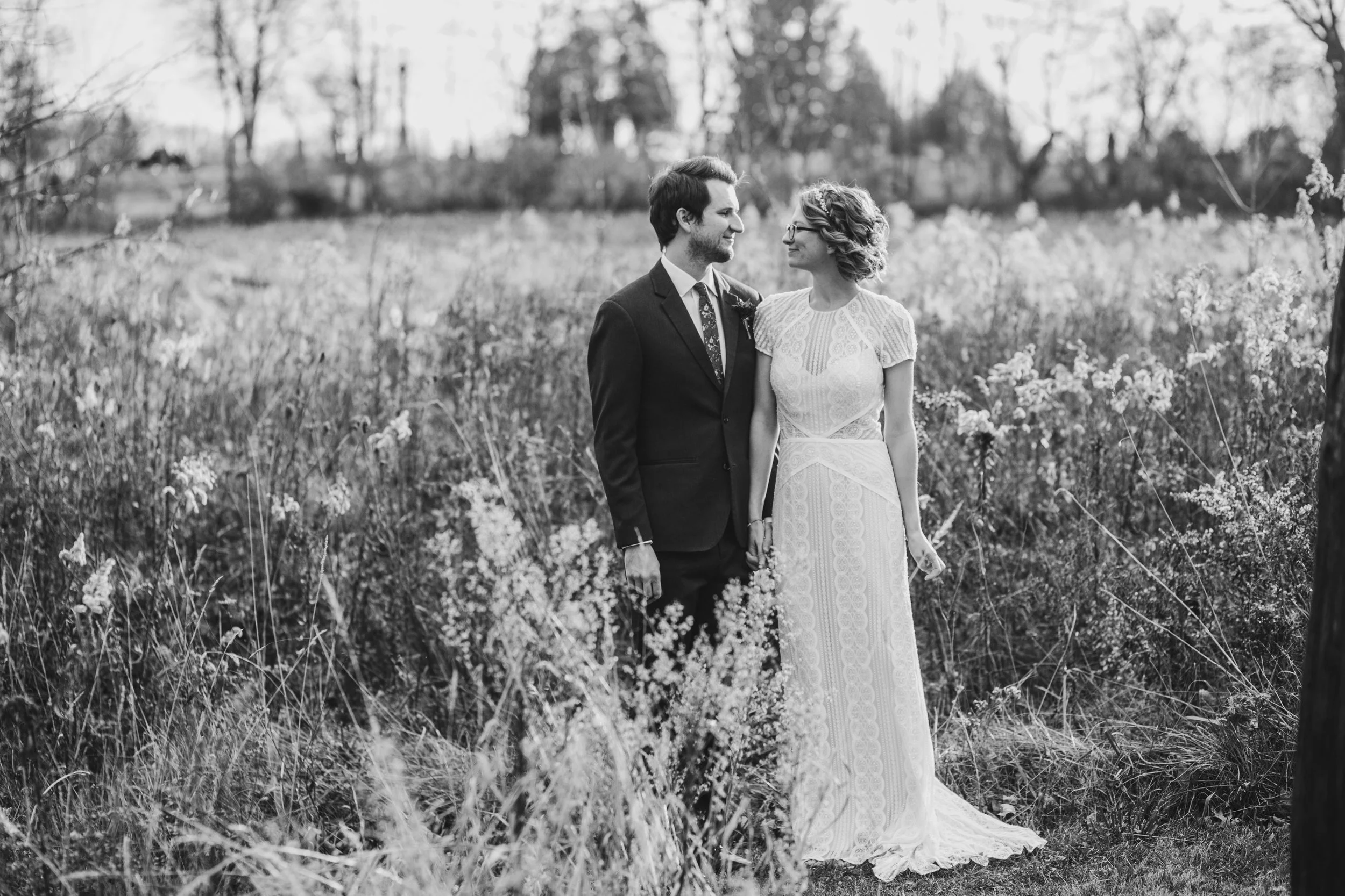 A couple dressed in wedding attire standing in a field of tall grass and flowers, holding hands and looking at each other.
