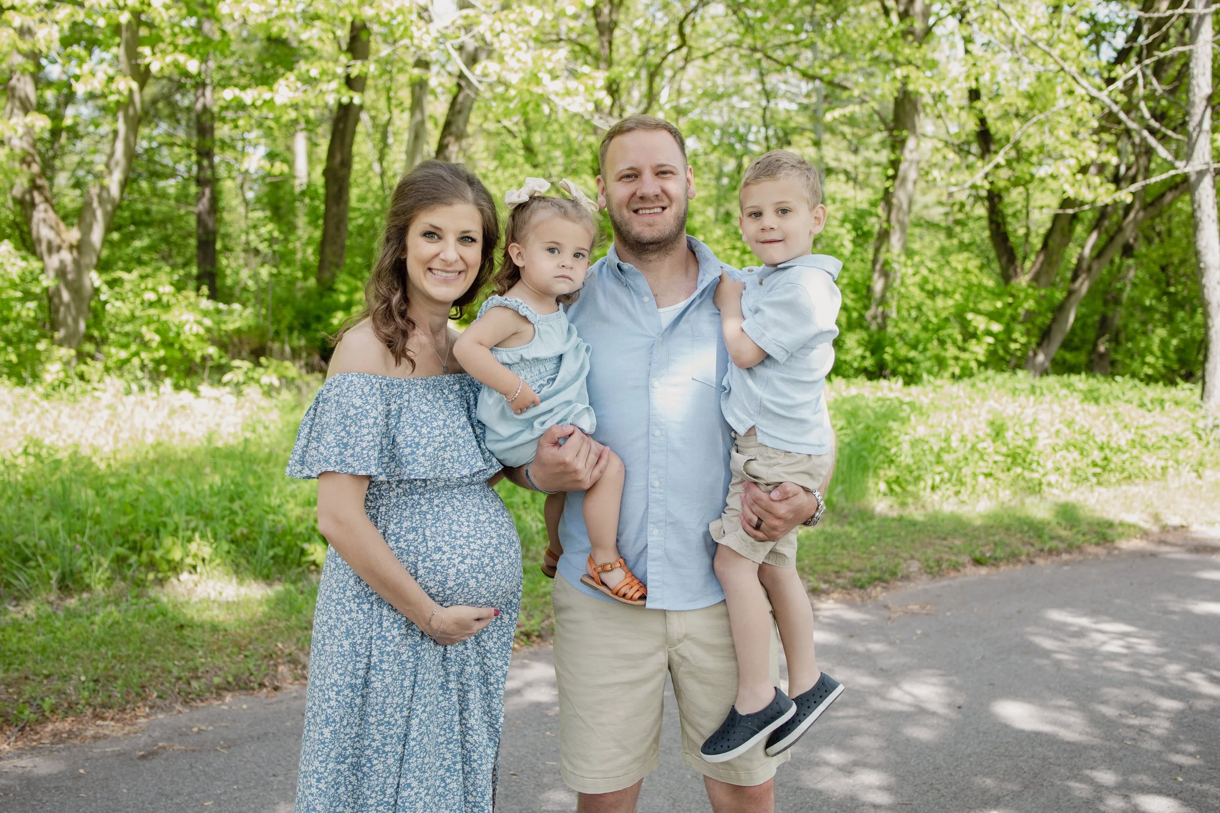 A family of four standing outdoors in a wooded park, smiling at the camera. The mother is on the left in a blue dress, the father is in the center in a blue shirt holding a young girl on his right arm, and a young boy is resting on his left arm. There are green trees and grass in the background.