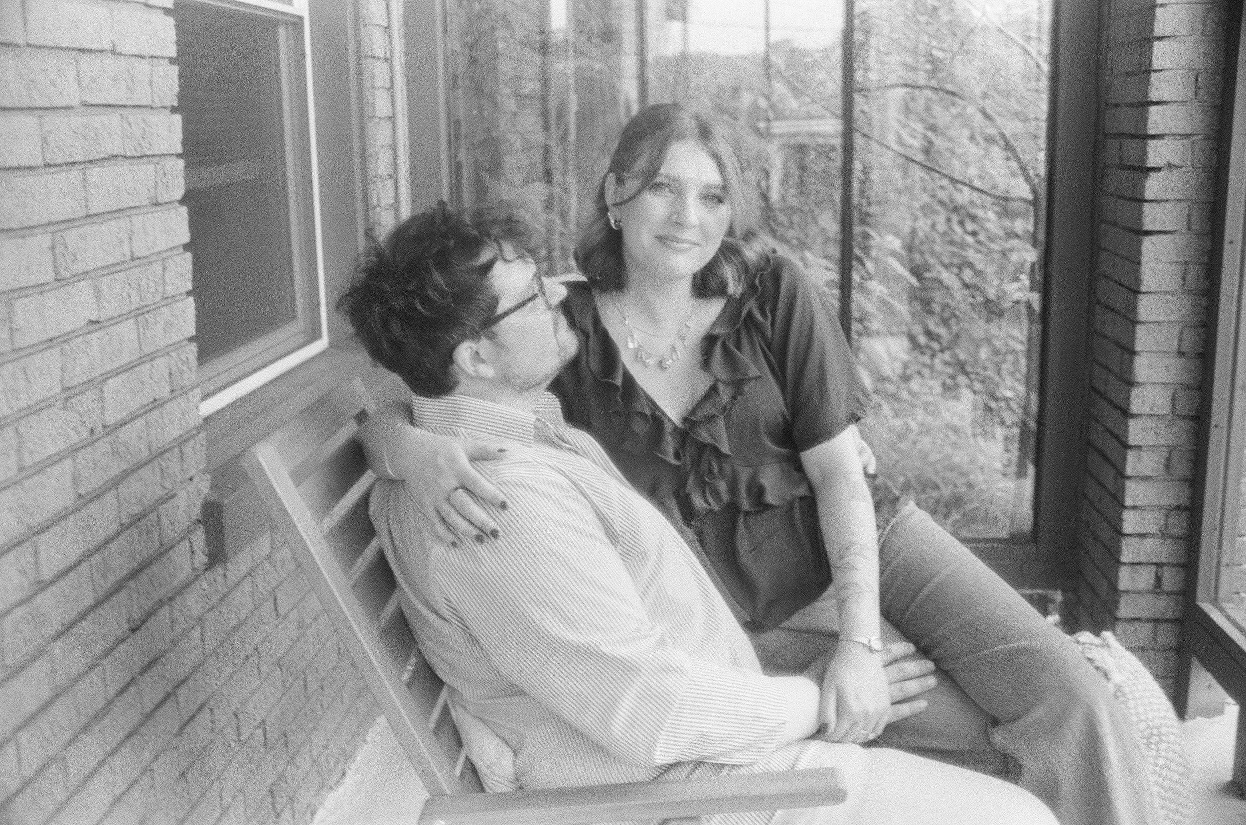 A couple sitting closely on a bench on a porch or balcony, with brick walls and large windows behind them, some trees outside, in black and white.