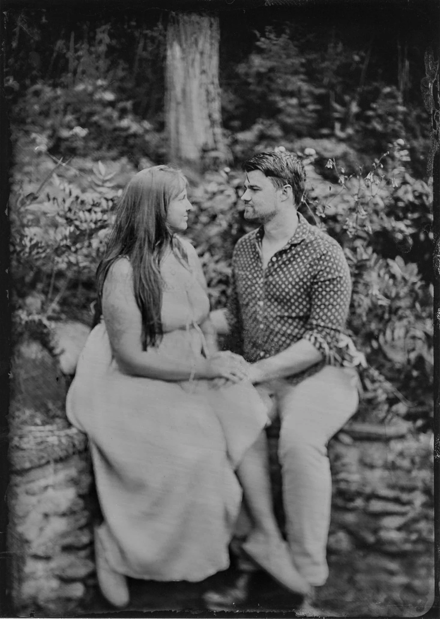 A black and white photo of a man and woman sitting outdoors on a bench, facing each other and holding hands, surrounded by trees and foliage.