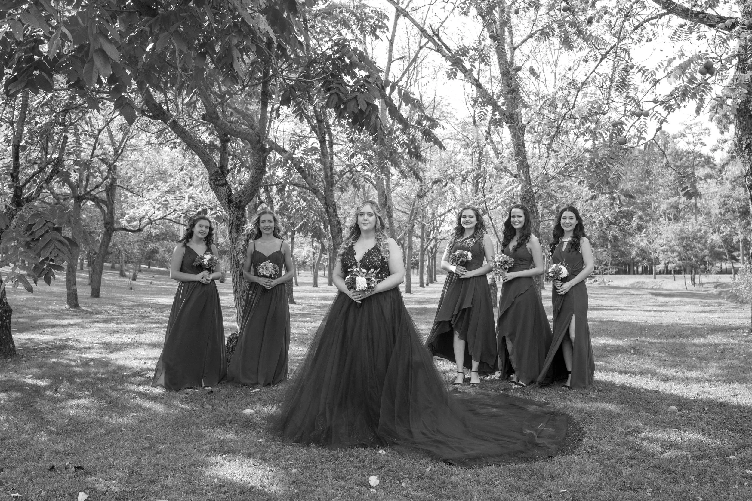 A group of six women dressed in formal gowns standing outdoors among trees, holding bouquets of flowers, in a natural setting with sunlight filtering through the trees.