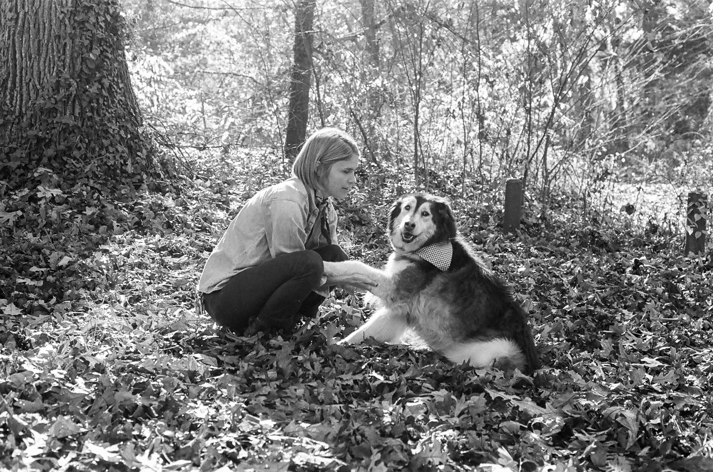 A woman with shoulder-length hair in a light jacket crouches next to a large dog with a thick coat and a bandana around its neck in a forest with fallen leaves and tall trees.