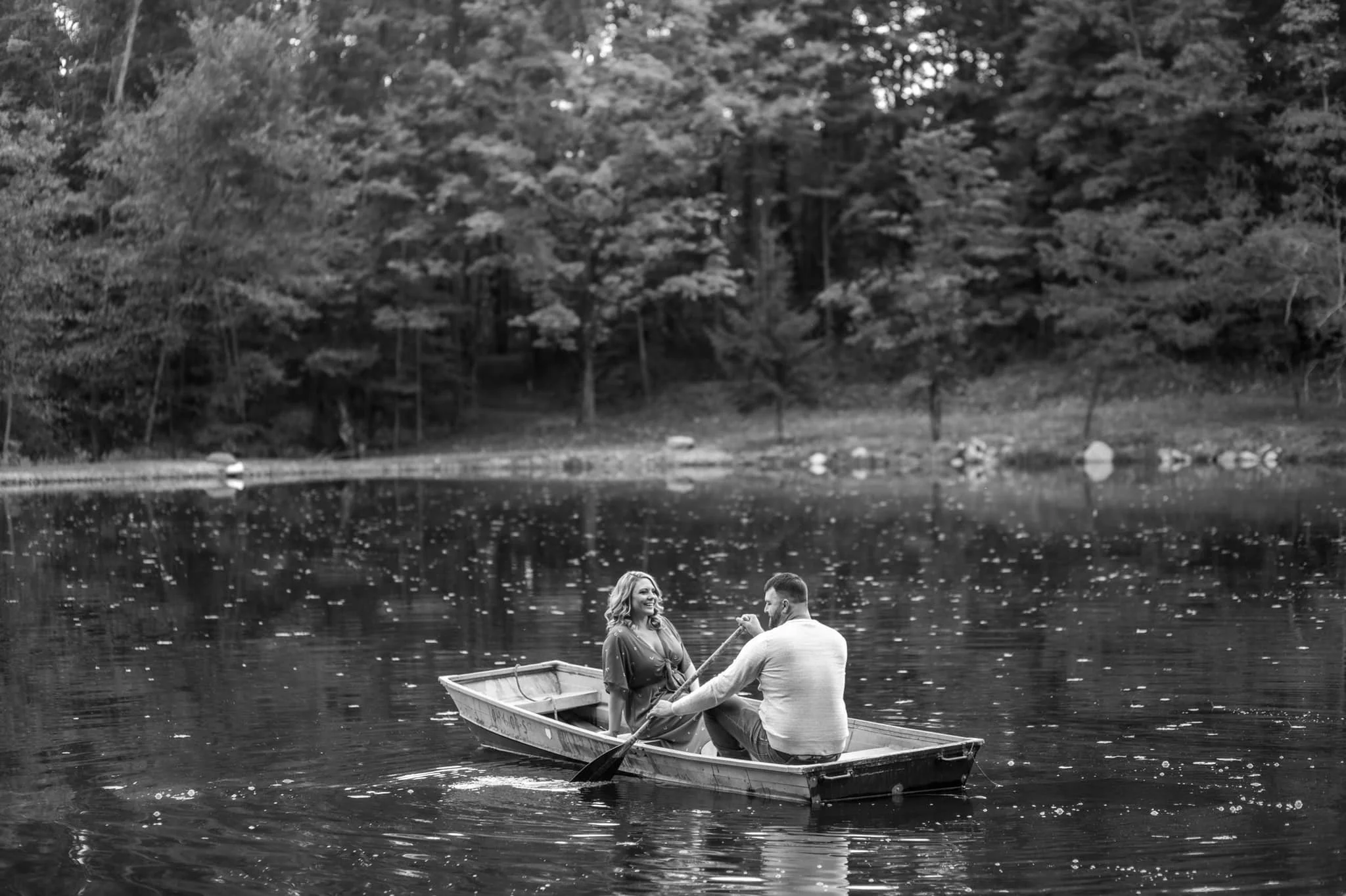A couple in a small boat on a lake, with the man rowing and the woman sitting and smiling, surrounded by a forested landscape in the background.