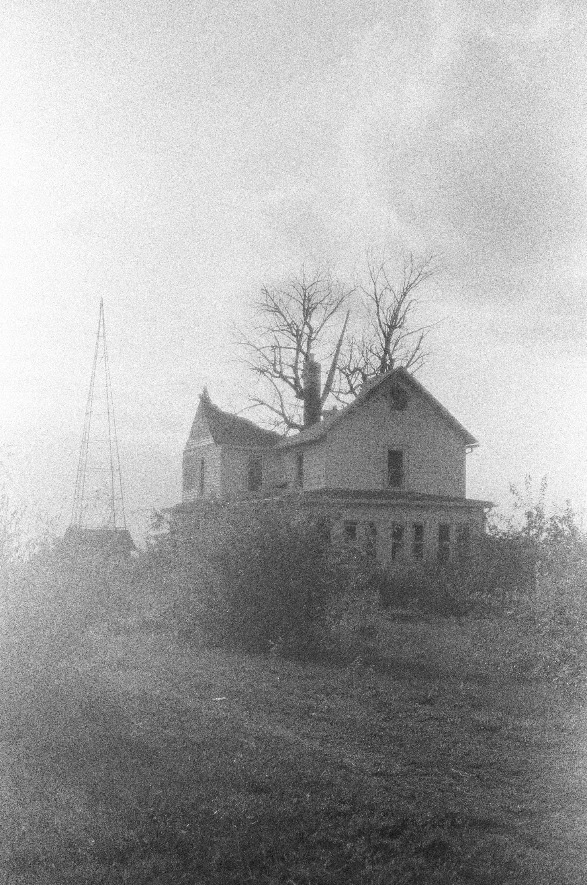 A black-and-white photo of an old house with a pointed roof and multiple stories, standing on a grassy plot with bushes in the foreground. A leafless tree is behind the house, and the sky appears cloudy and overcast.