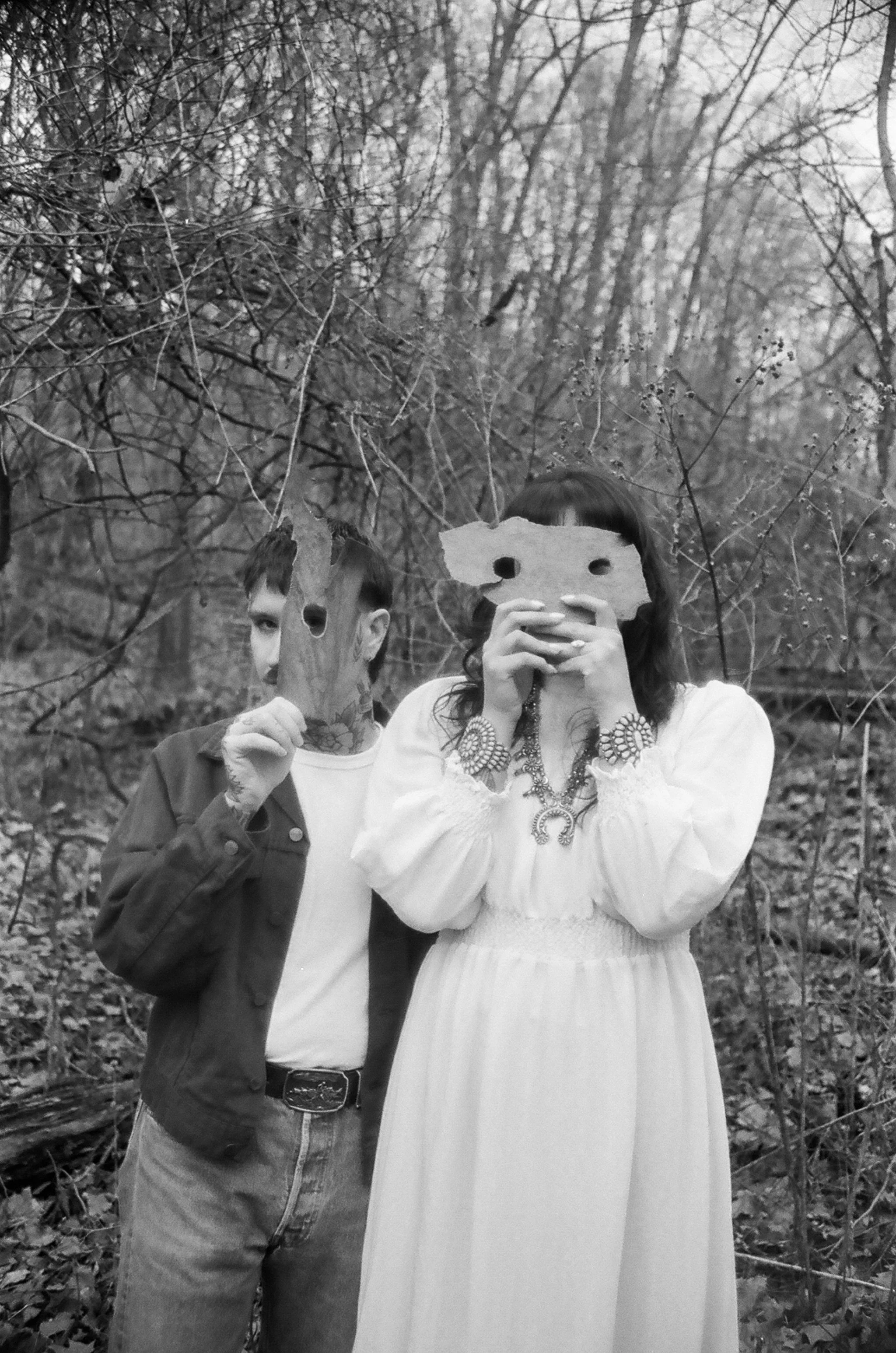 Black and white photo of a man and a woman showing faces through cutouts in masks made of paper, standing outdoors among barren trees and fallen leaves, taking a selfie.