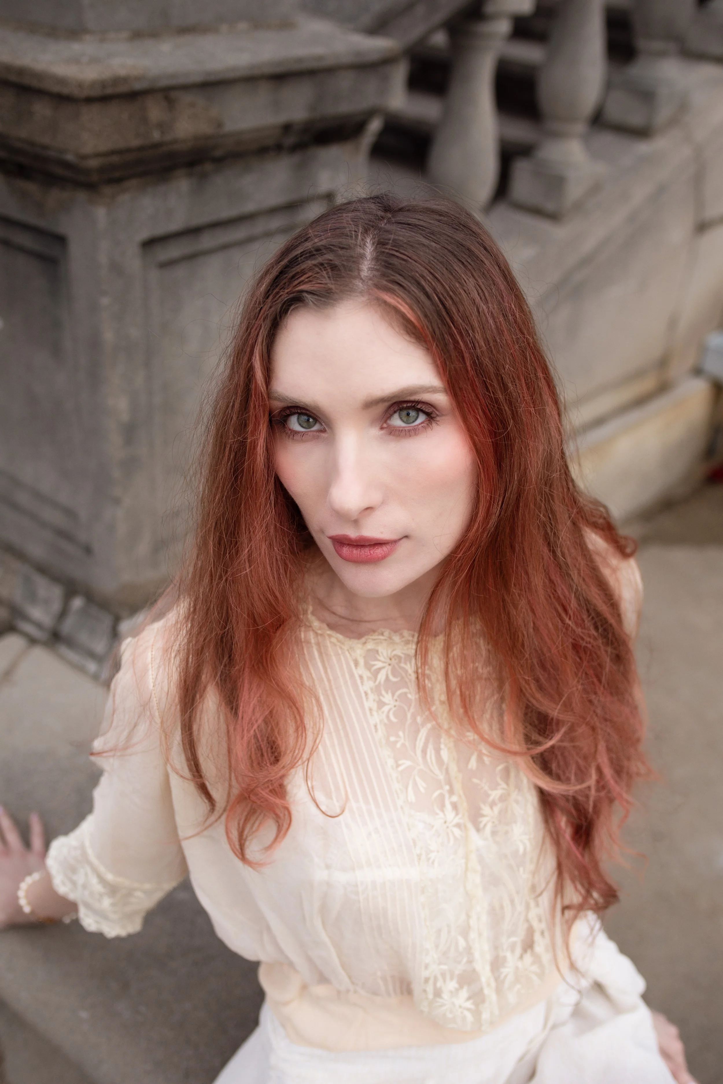 A woman with long reddish hair and gray eyes is looking up at the camera with a soft expression. She is wearing a cream-colored lace dress and standing outdoors against a stone balustrade.