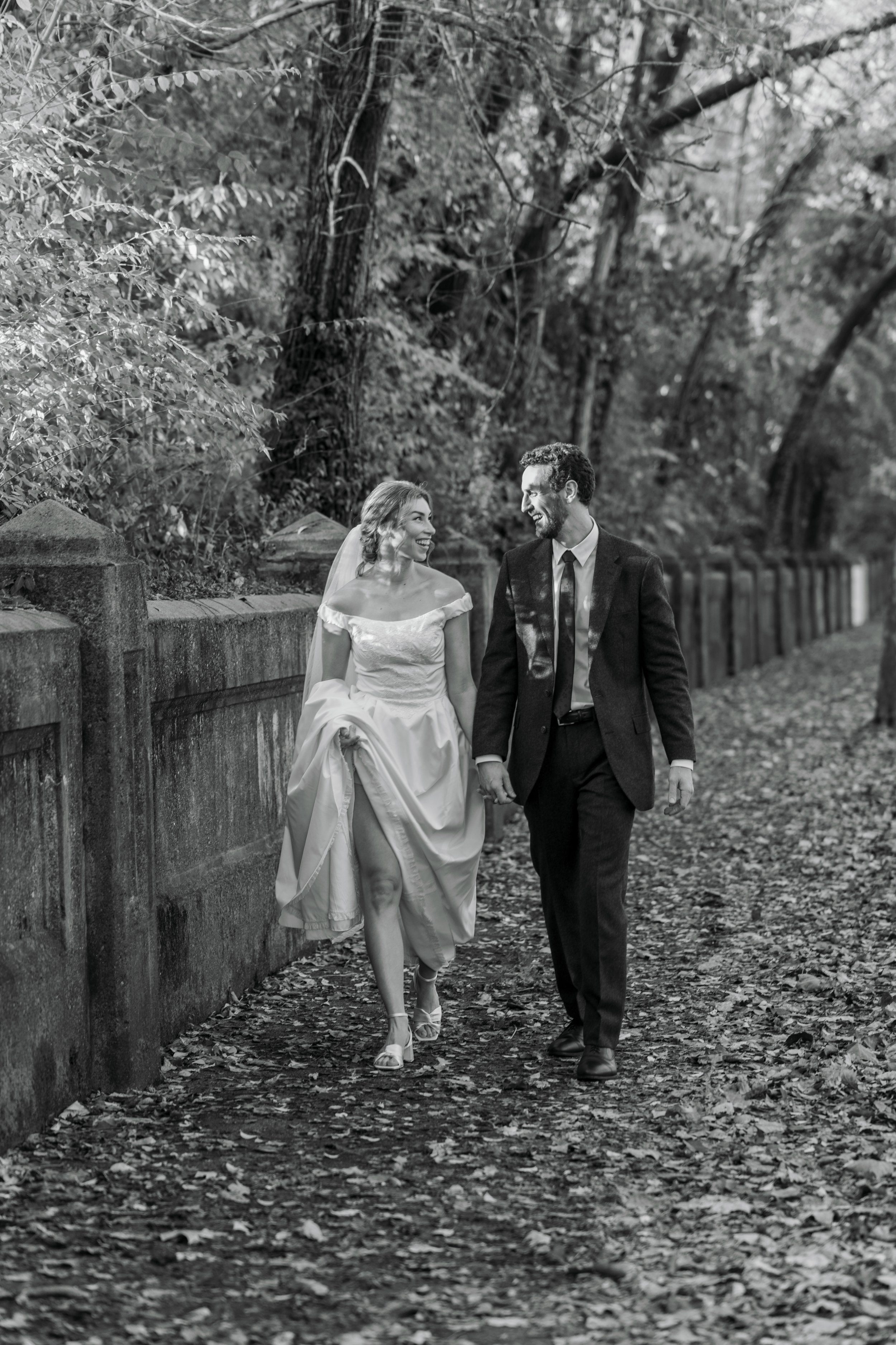 A black and white photo of a newlywed couple walking hand in hand on a tree-lined path with fallen leaves, the woman in a wedding dress and the man in a suit, smiling and looking at each other.