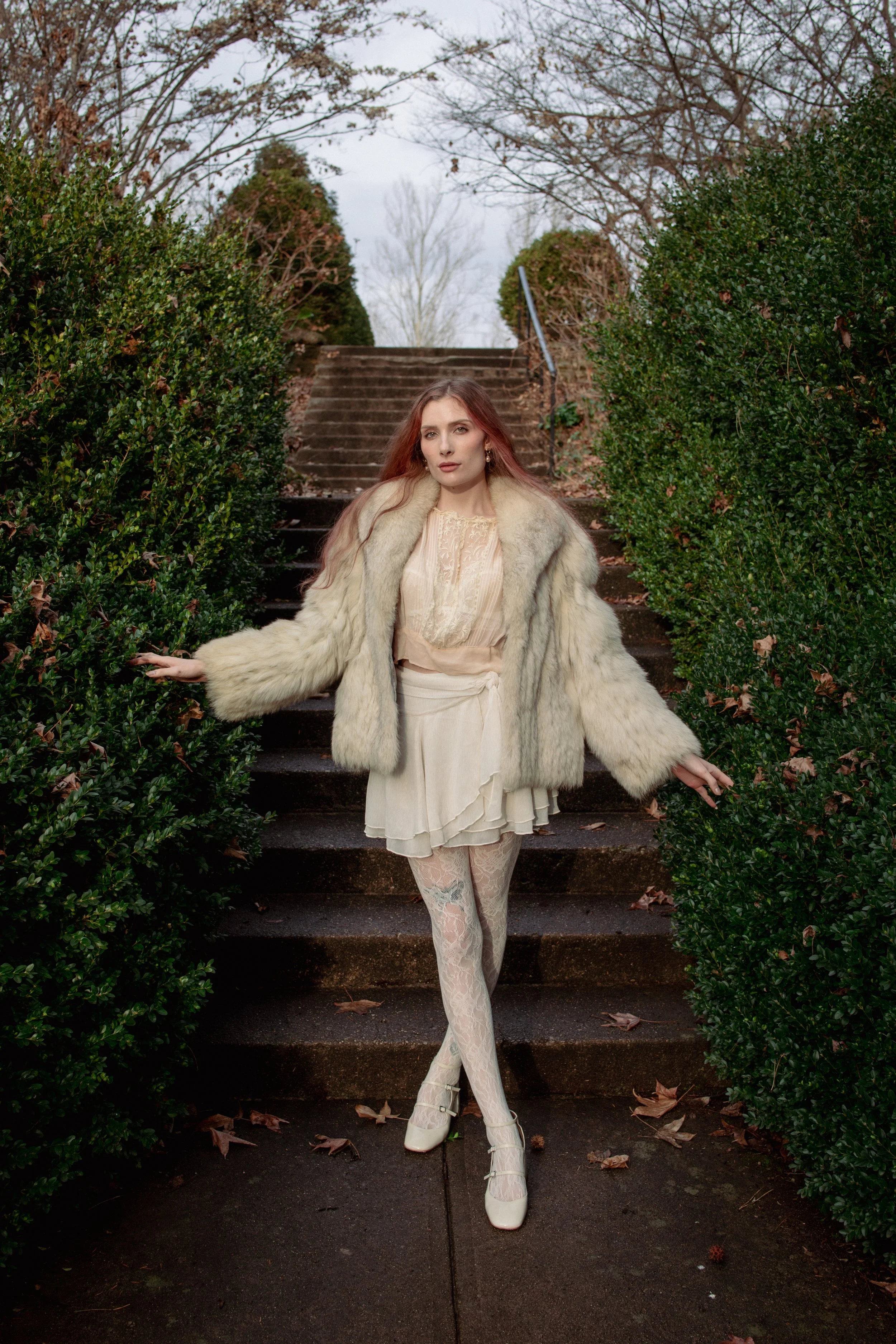 A woman with long reddish hair standing on outdoor stairs, wearing a cream-colored dress, lace tights, white heels, and a large fur coat, with bushes on either side of the stairs and trees in the background.