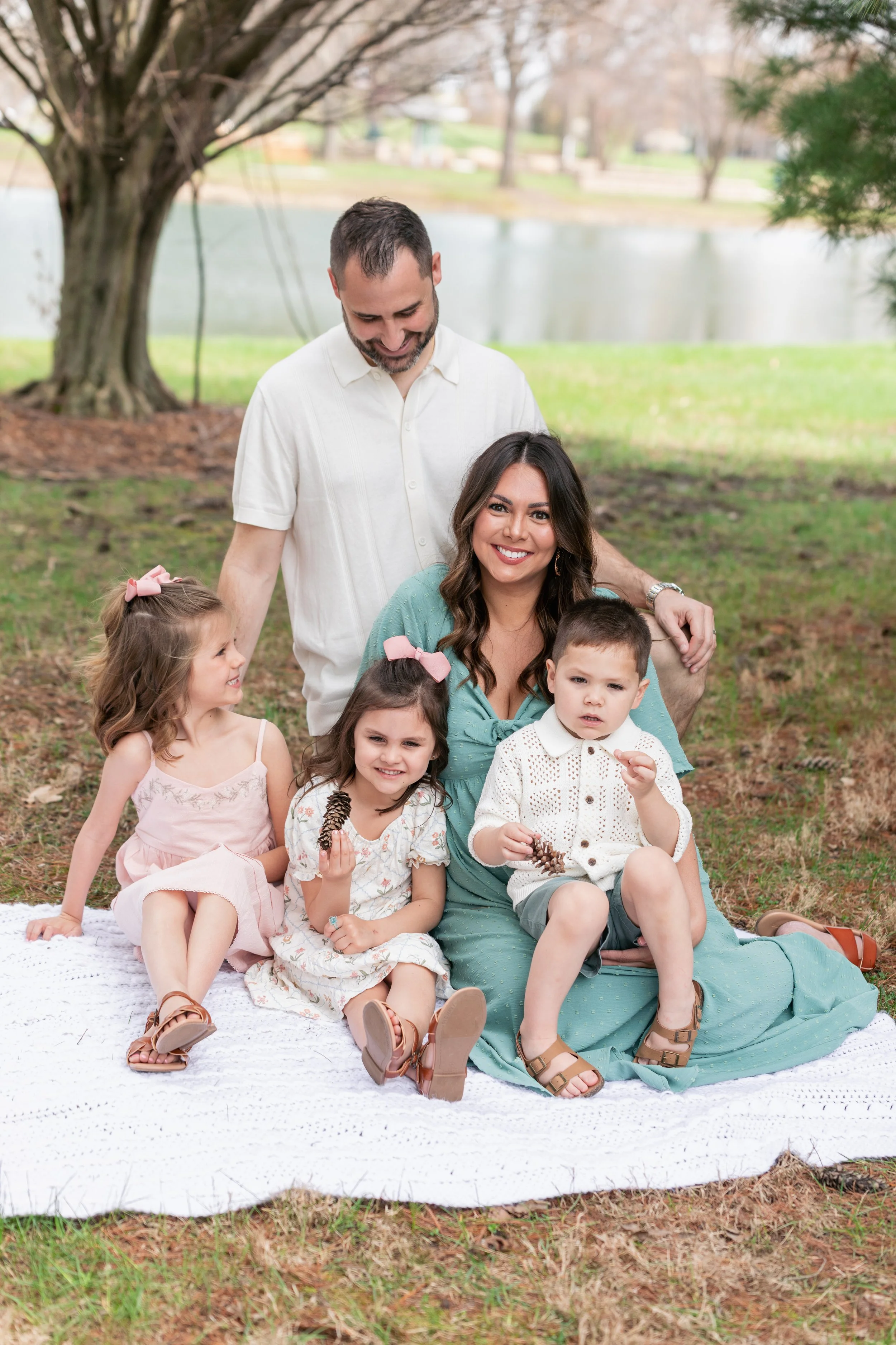 A smiling woman sitting on a blanket outdoors with children, surrounded by trees and a lake.