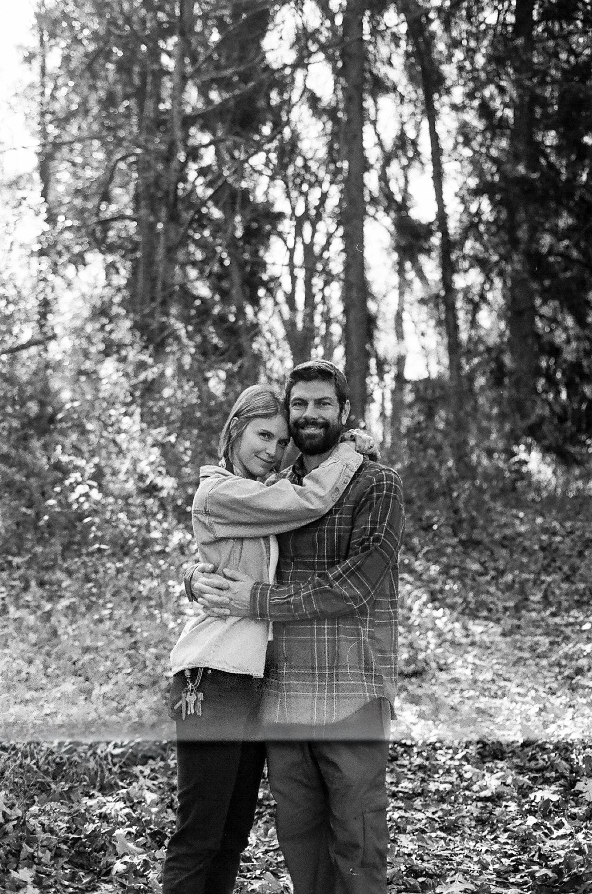 A black and white photo of a man and a woman standing in a forest, embracing each other and smiling at the camera.