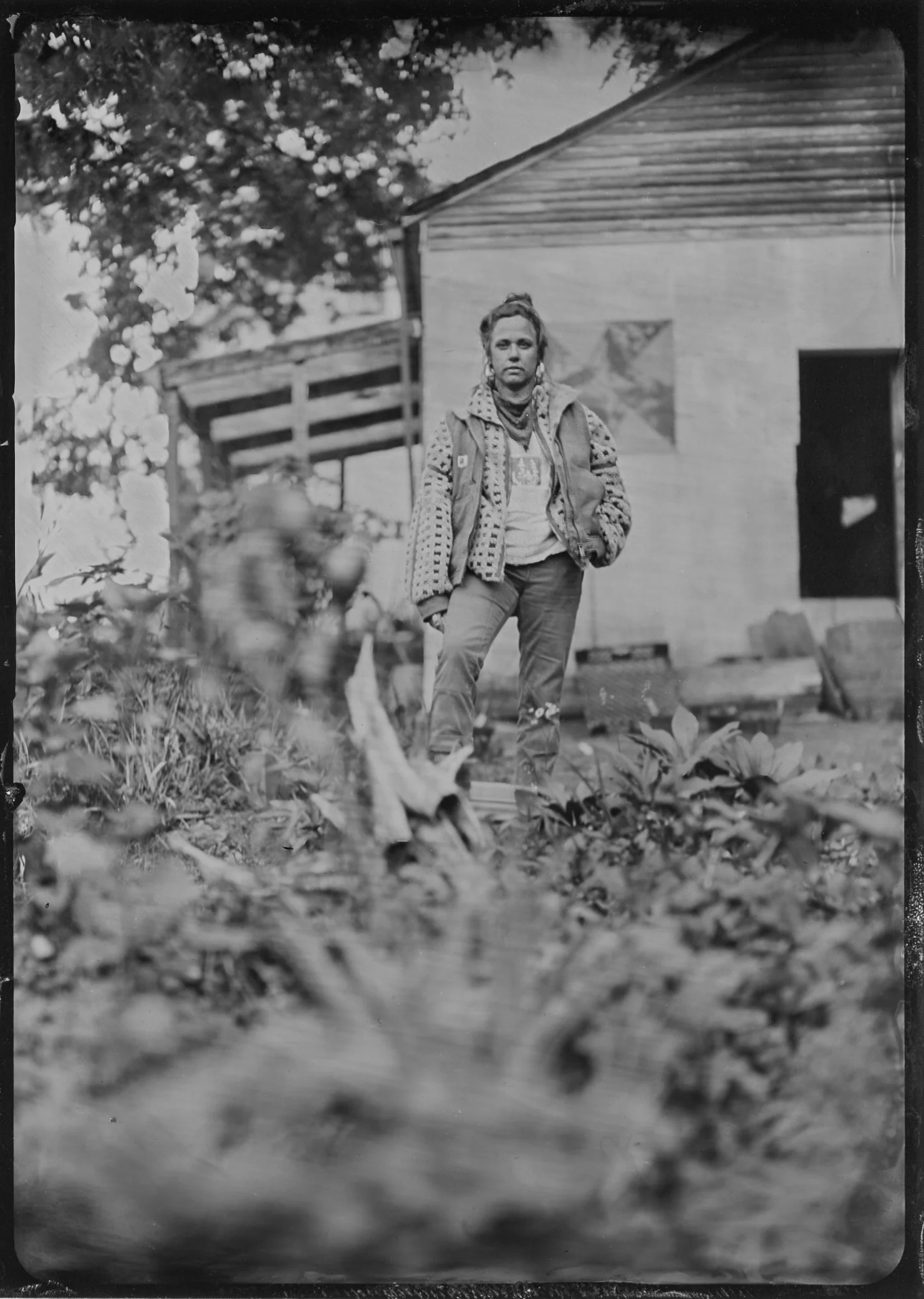 A woman standing outdoors in front of a wooden building, wearing casual clothes and a patterned jacket, with plants and trees in the foreground.
