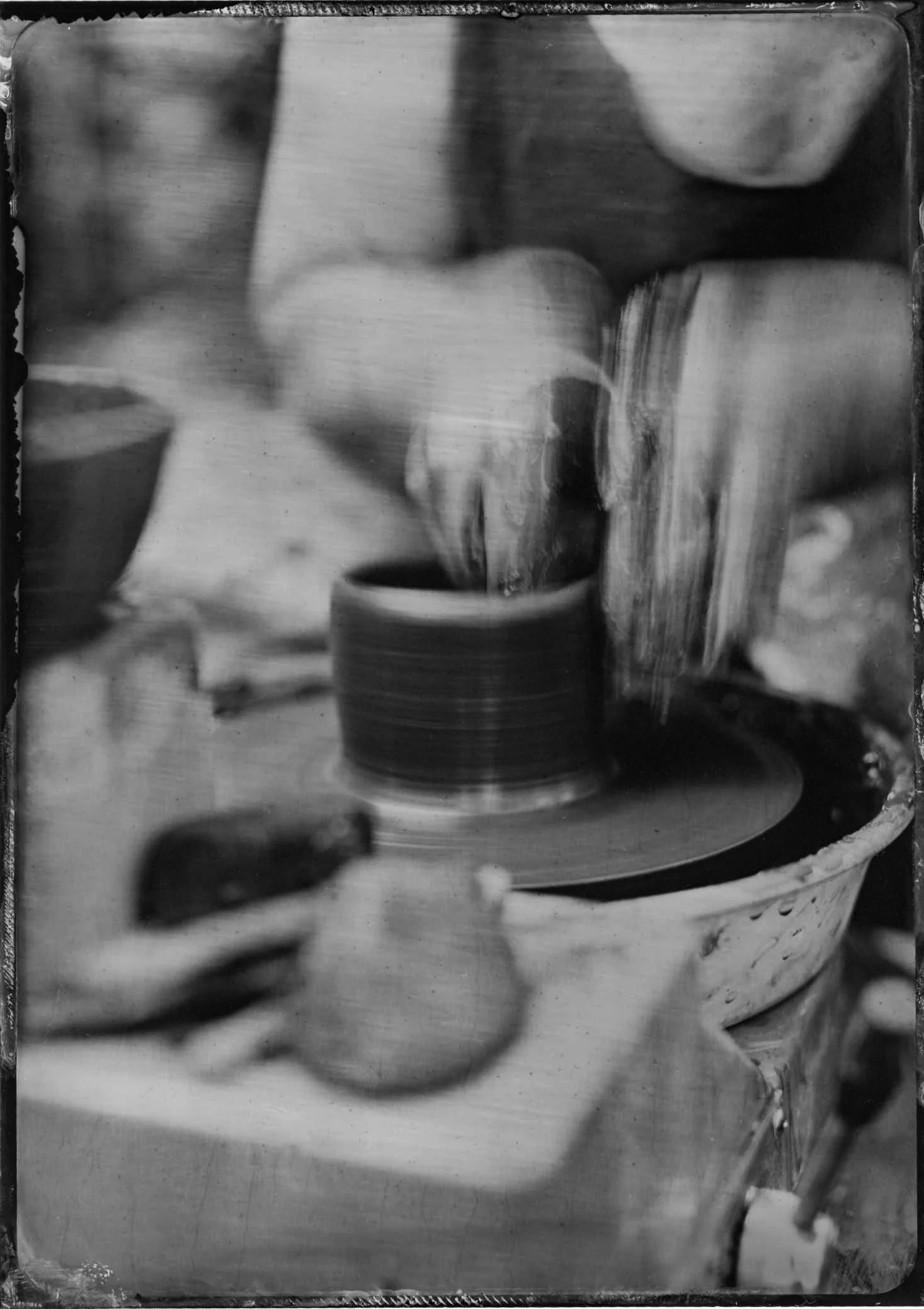 Black and white photograph of a cup with a hot beverage, with steam rising from it, placed on a table. There is a spoon next to the cup and a container or pitcher in the background.