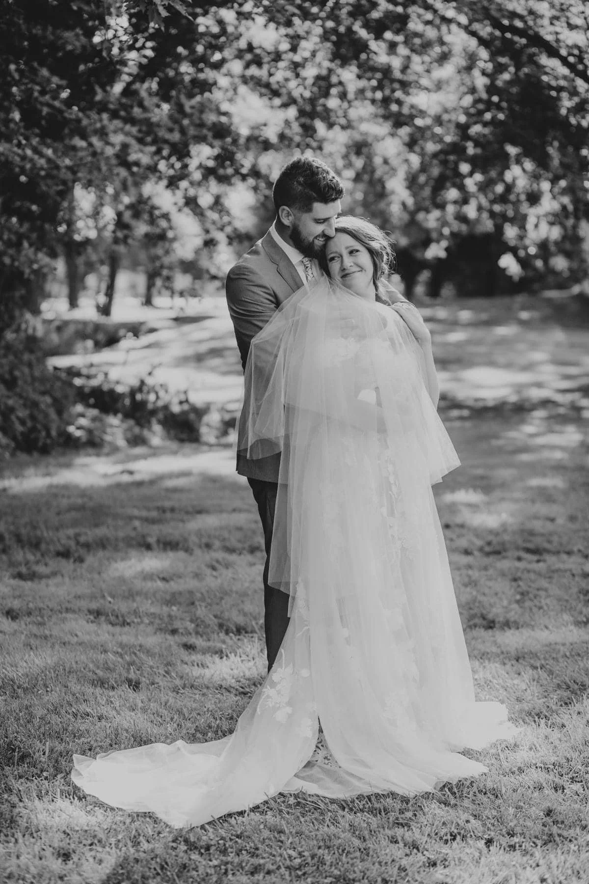 A black-and-white photograph of a bride and groom embracing outdoors on a grassy area with trees in the background. The bride is wearing a long wedding dress with a train and veil, and the groom is in a suit. They are smiling, with the groom leaning 
