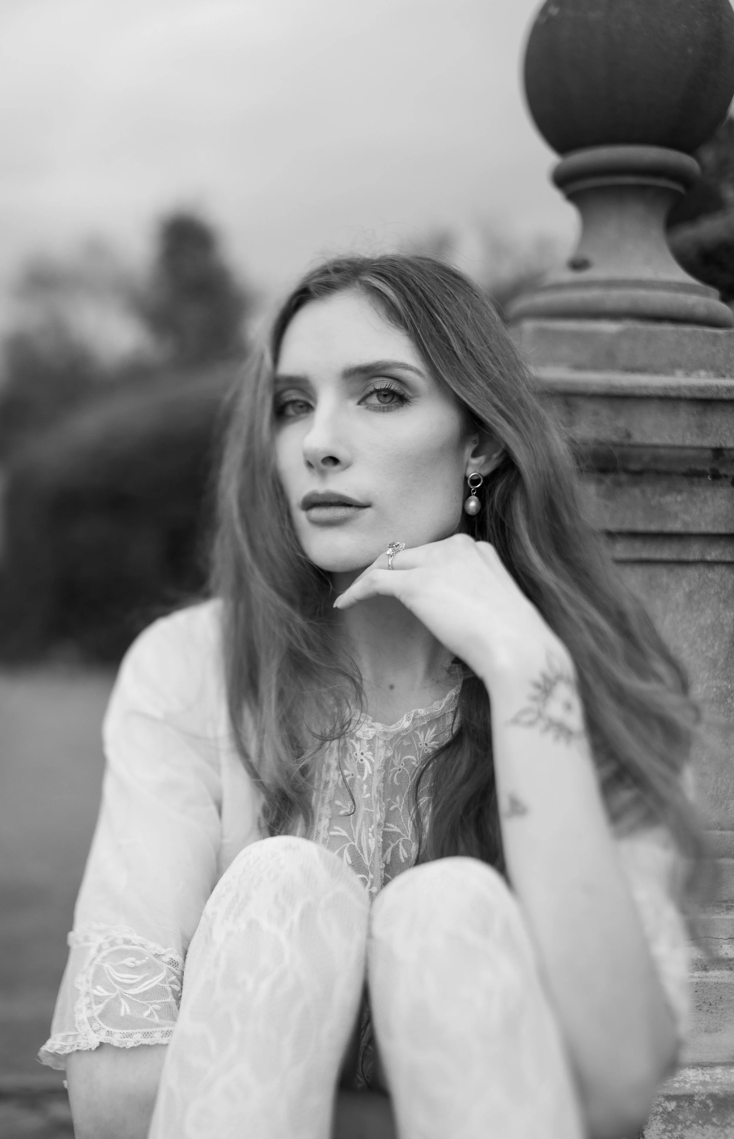 A black and white photograph of a woman with long hair sitting outdoors near a decorative stone fence. She is looking at the camera with her hand resting near her chin, wearing earrings and a lace top.