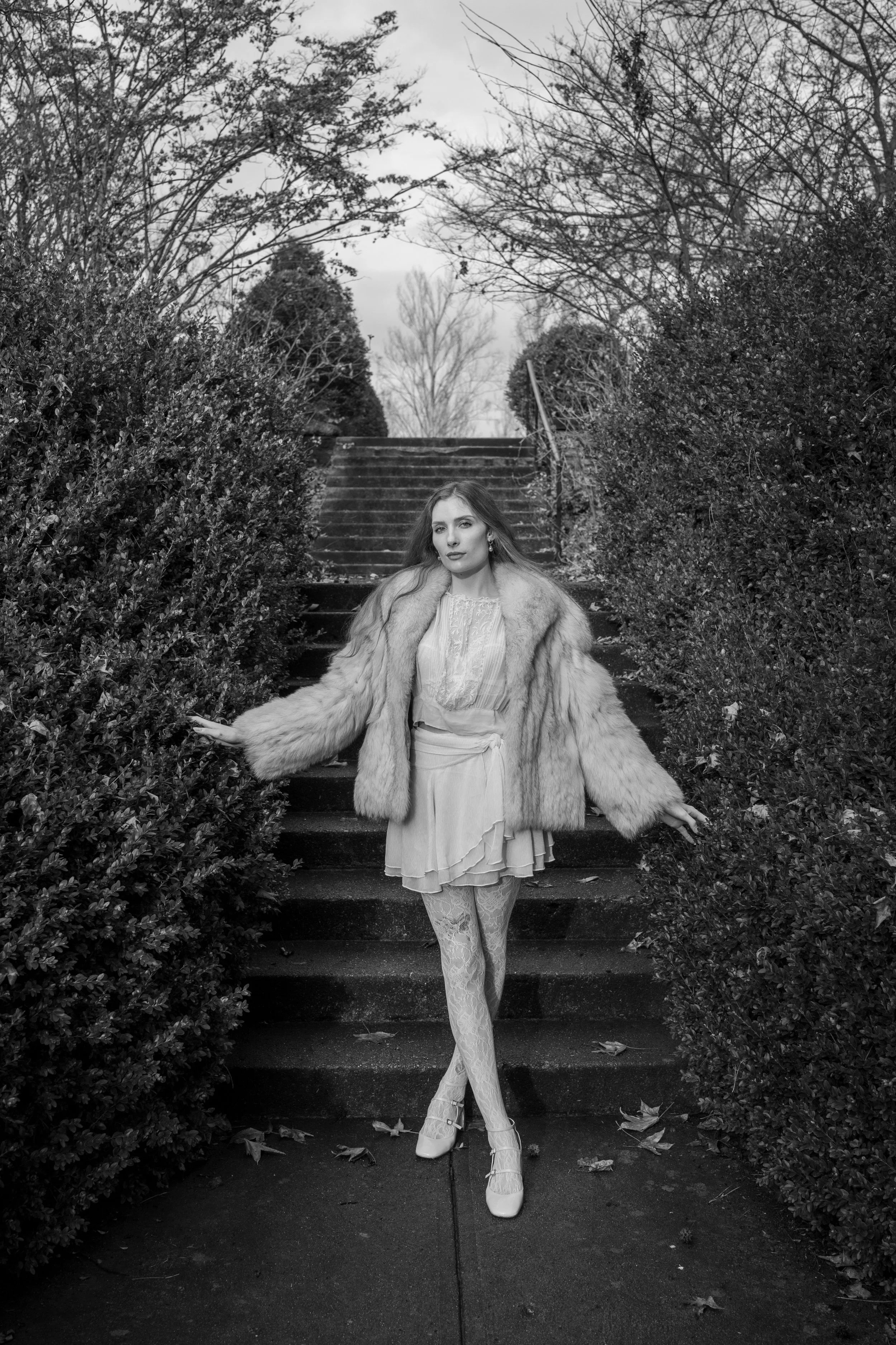 A woman with long hair in a vintage dress and high heels, wearing a fur coat, standing on outdoor stairs surrounded by bushes and trees, with a cloudy sky overhead.
