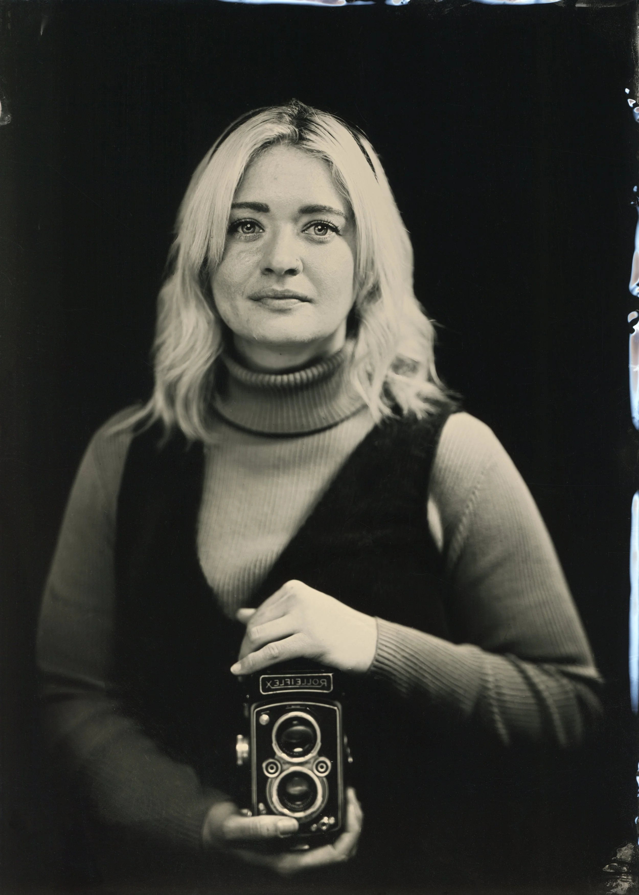 Black and white photo of a woman with blonde hair taking a mirror selfie with a vintage Rolleiflex camera. She is wearing a turtleneck sweater and has a small nose piercing.