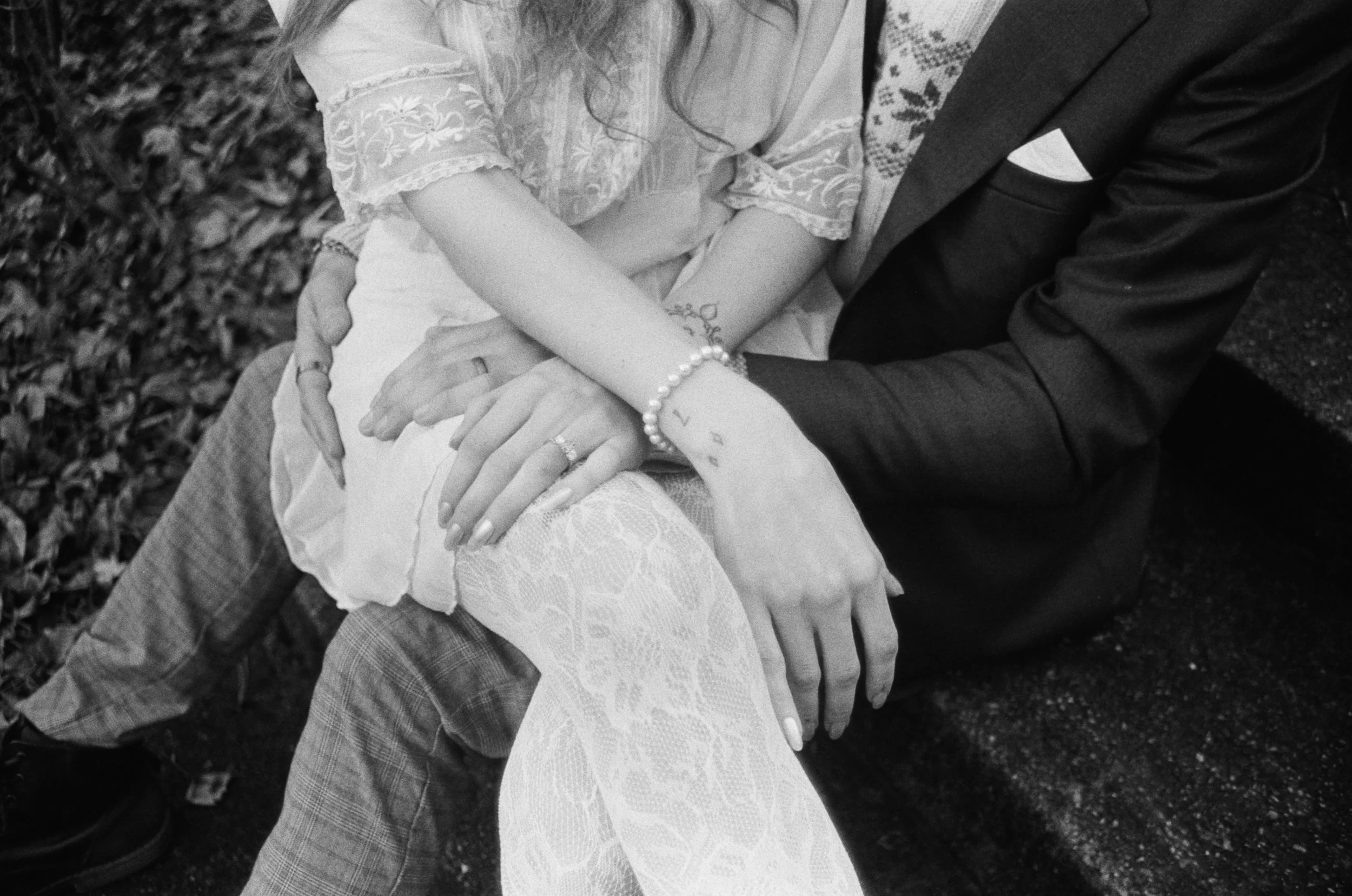 Close-up of several hands intertwined, showing wedding rings and jewelry, with part of a woman's lace dress and a man's suit visible; background of fallen leaves.