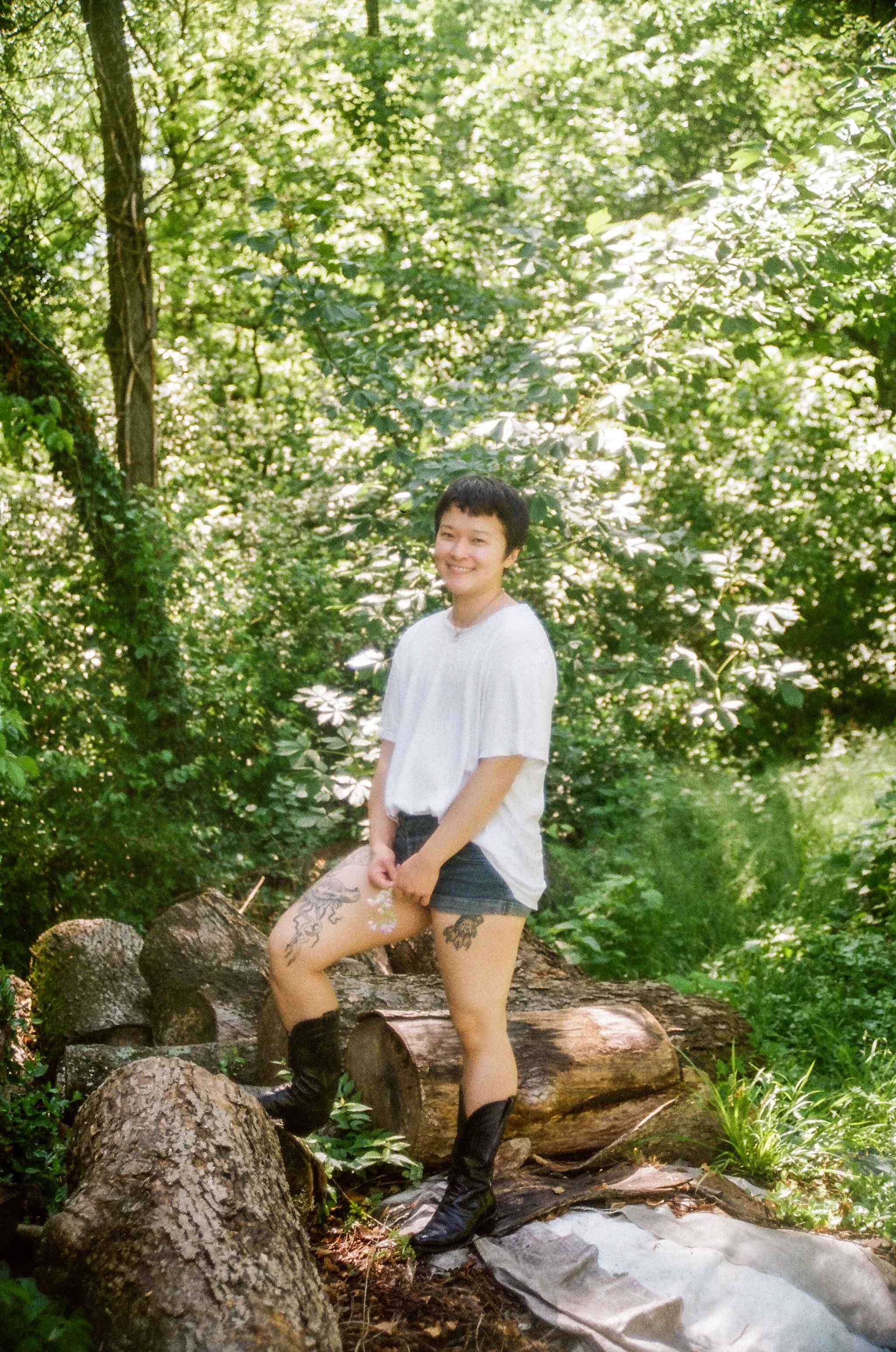 A young woman with tattoos on her thigh and wearing a white t-shirt, black shorts, and black boots, standing outdoors on logs in a wooded area with green foliage.