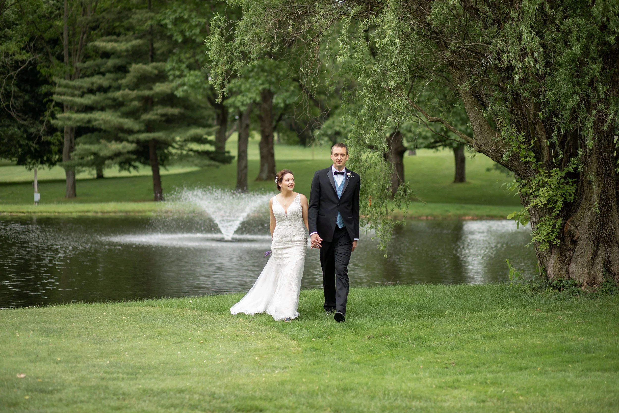 A bride and groom holding hands walking by a pond in a park with green trees.