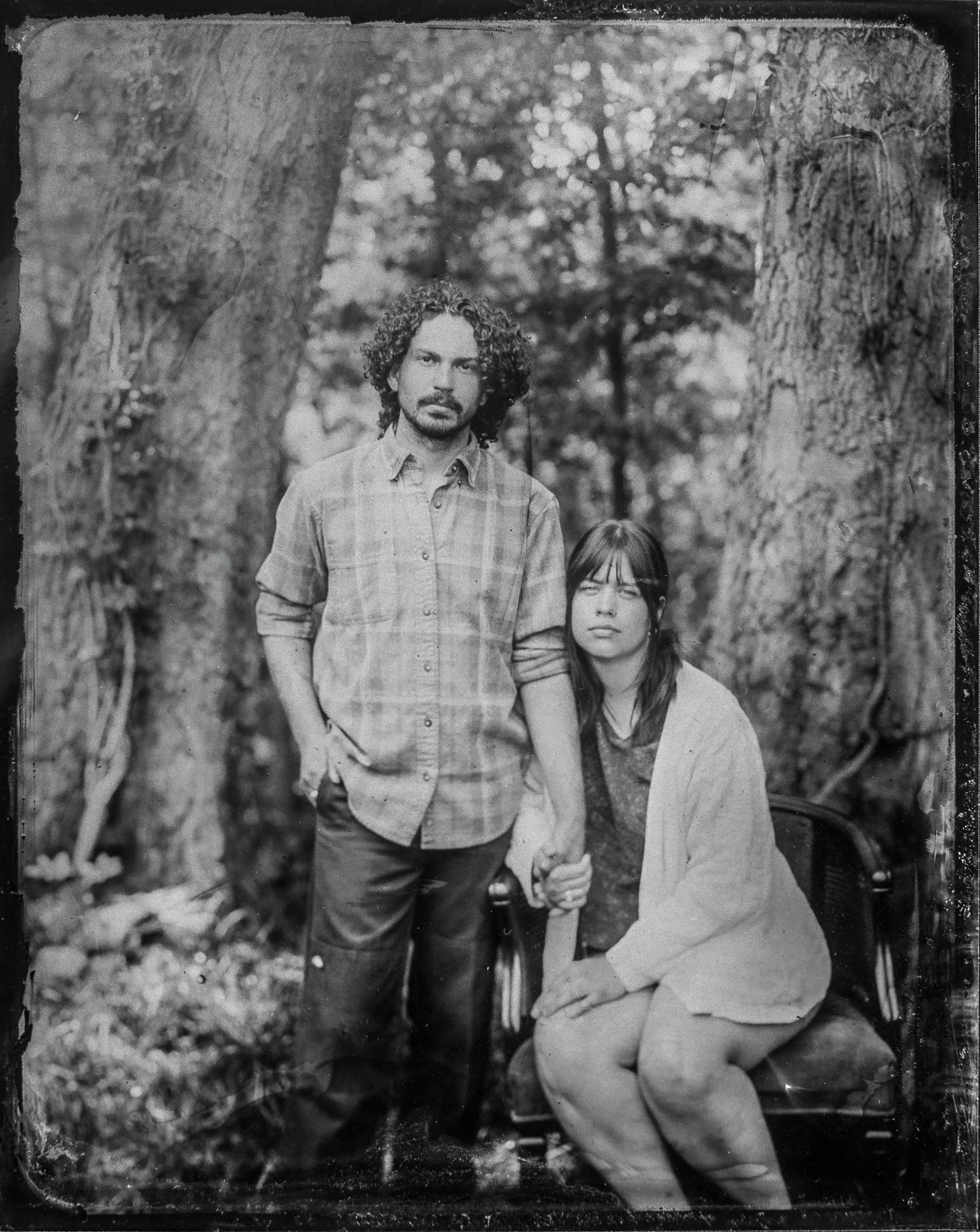 Black and white photograph of a man and a woman outdoors, with large tree trunks in the background. The man has curly hair, a beard, and is wearing a plaid shirt with the sleeves rolled up. The woman has straight hair, glasses, and is wearing a light-colored jacket or sweater and is sitting in a wooden chair. The man is standing with his right hand in his pocket, and the woman is seated with her hands on her thighs.