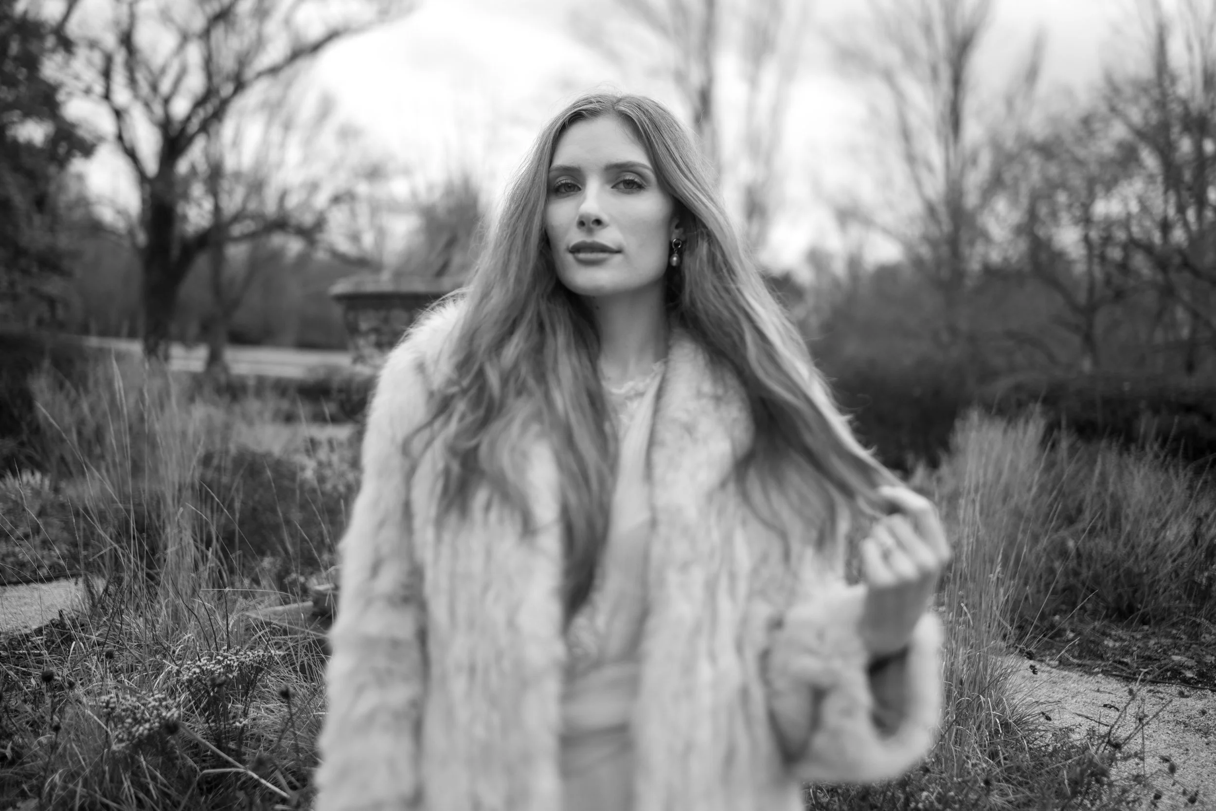 Black and white photo of a woman with long hair and earrings, outdoors in a natural setting with trees and grass, looking directly at the camera.