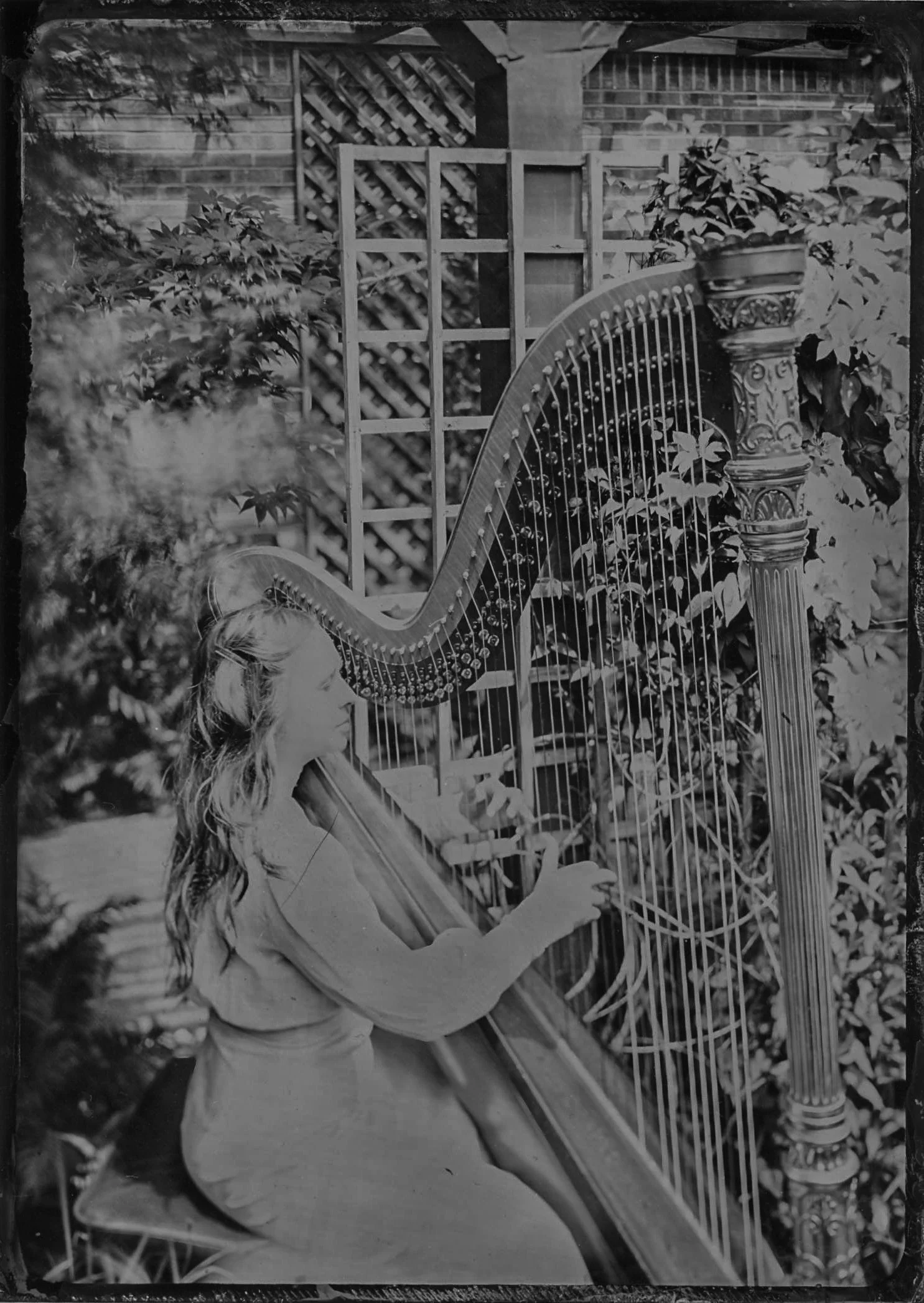 A young girl with long hair sitting outdoors playing a large harp among trees and plants.