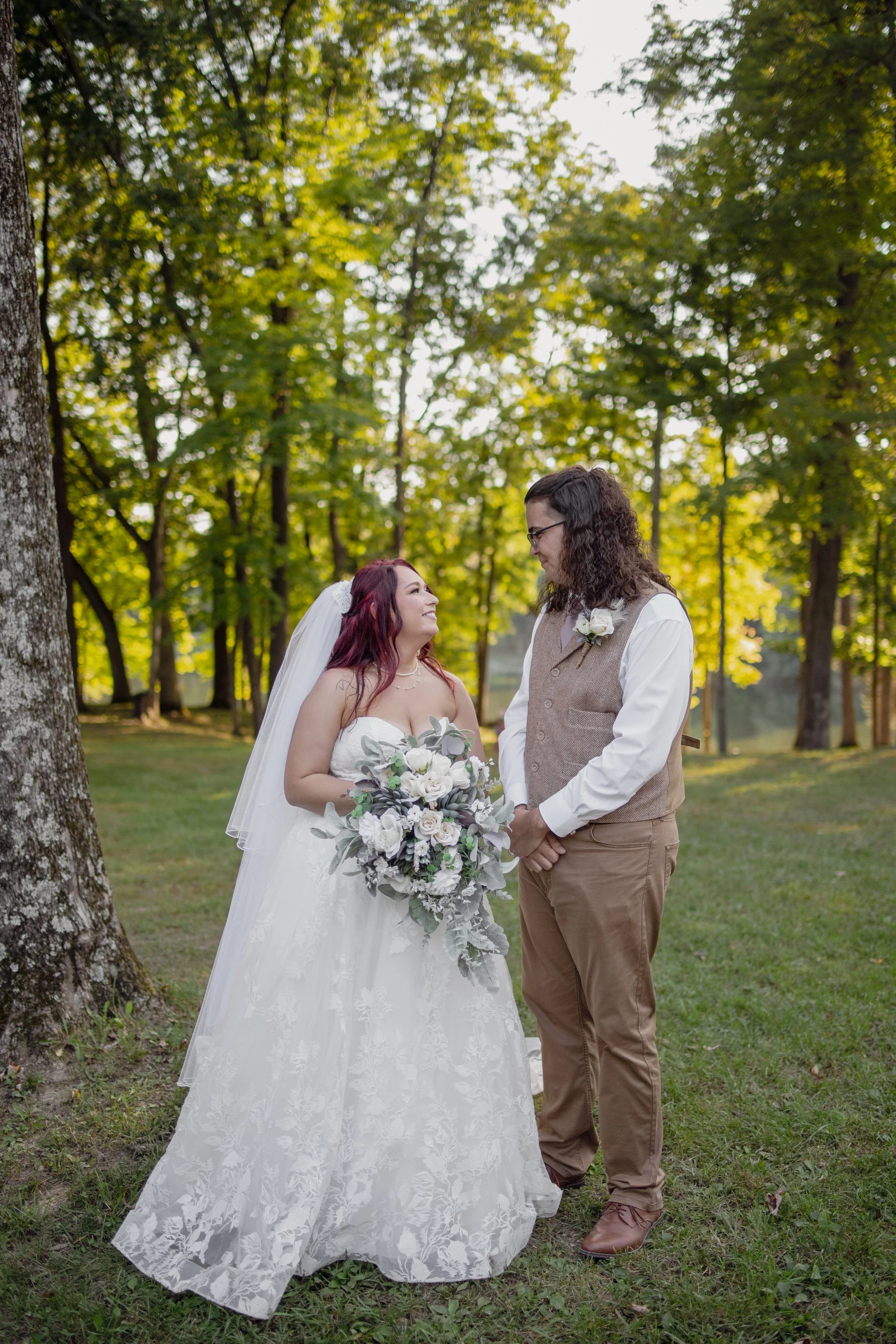 A bride and groom standing outdoors in a park with green trees in the background, holding hands and gazing at each other. The bride is wearing a white wedding gown with a veil and holding a bouquet of white flowers, while the groom is wearing a beige