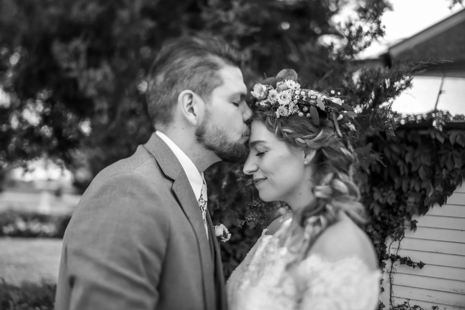 Black and white photo of a newlywed couple, with the groom kissing the bride's forehead as she smiles with her eyes closed, outdoors with trees and a house in the background.