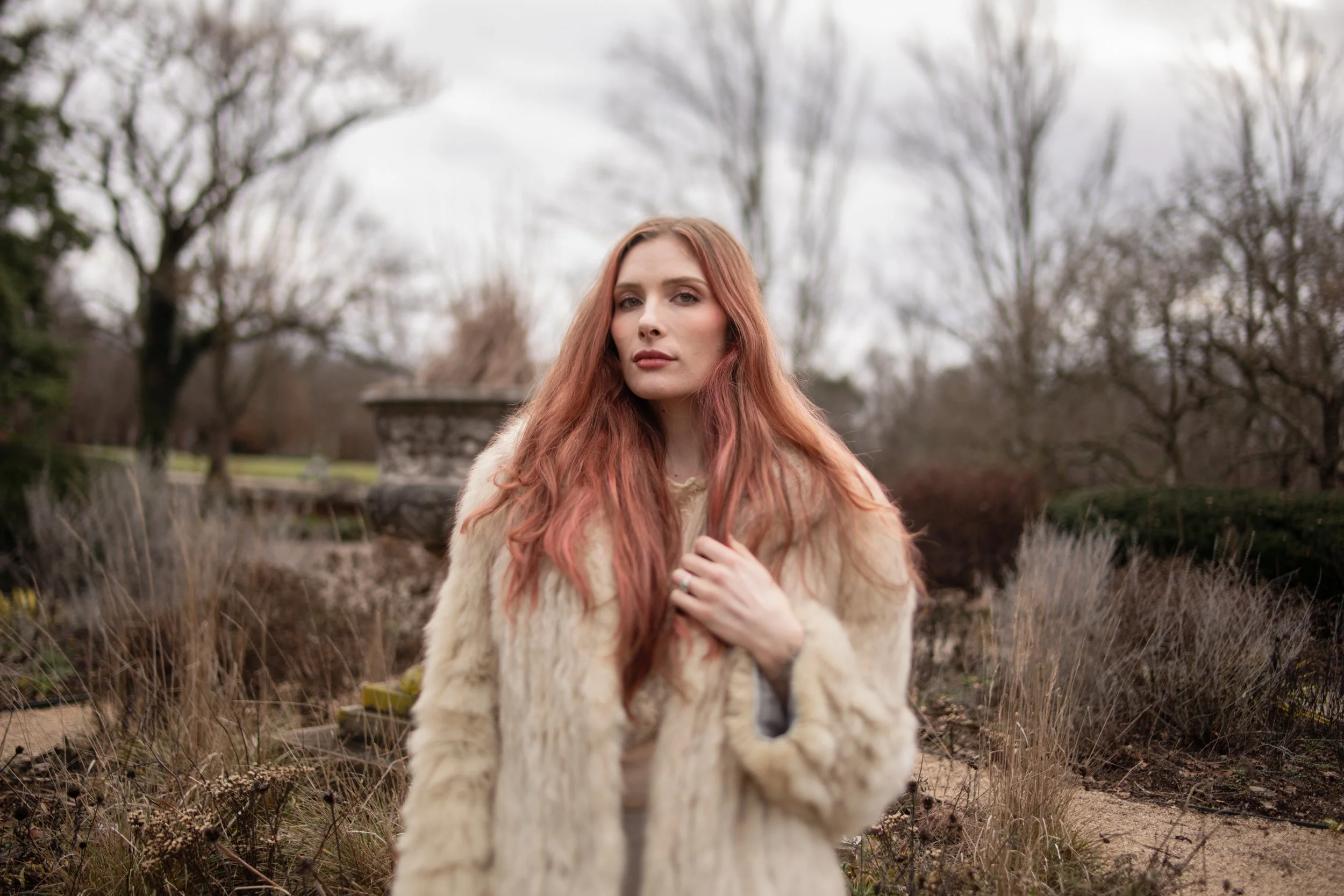 A woman with long red hair and fair skin standing outdoors in a garden or park with leafless trees in the background, wearing a beige or light-colored fur coat, gazing into the camera.