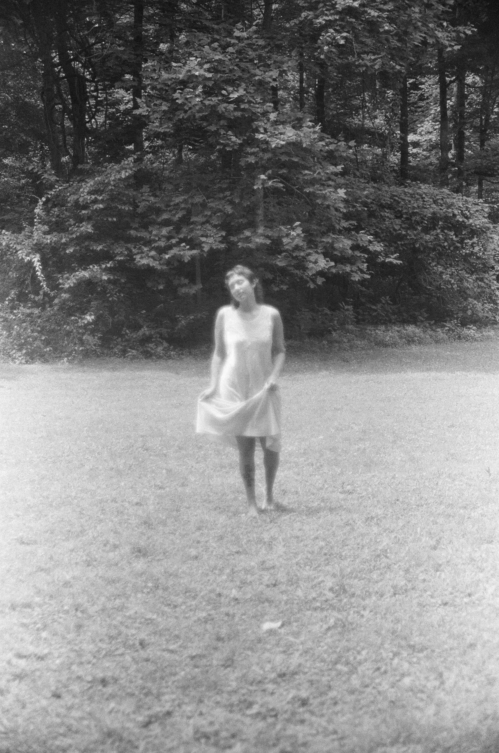 A woman in a dress standing on grass with a backdrop of trees.
