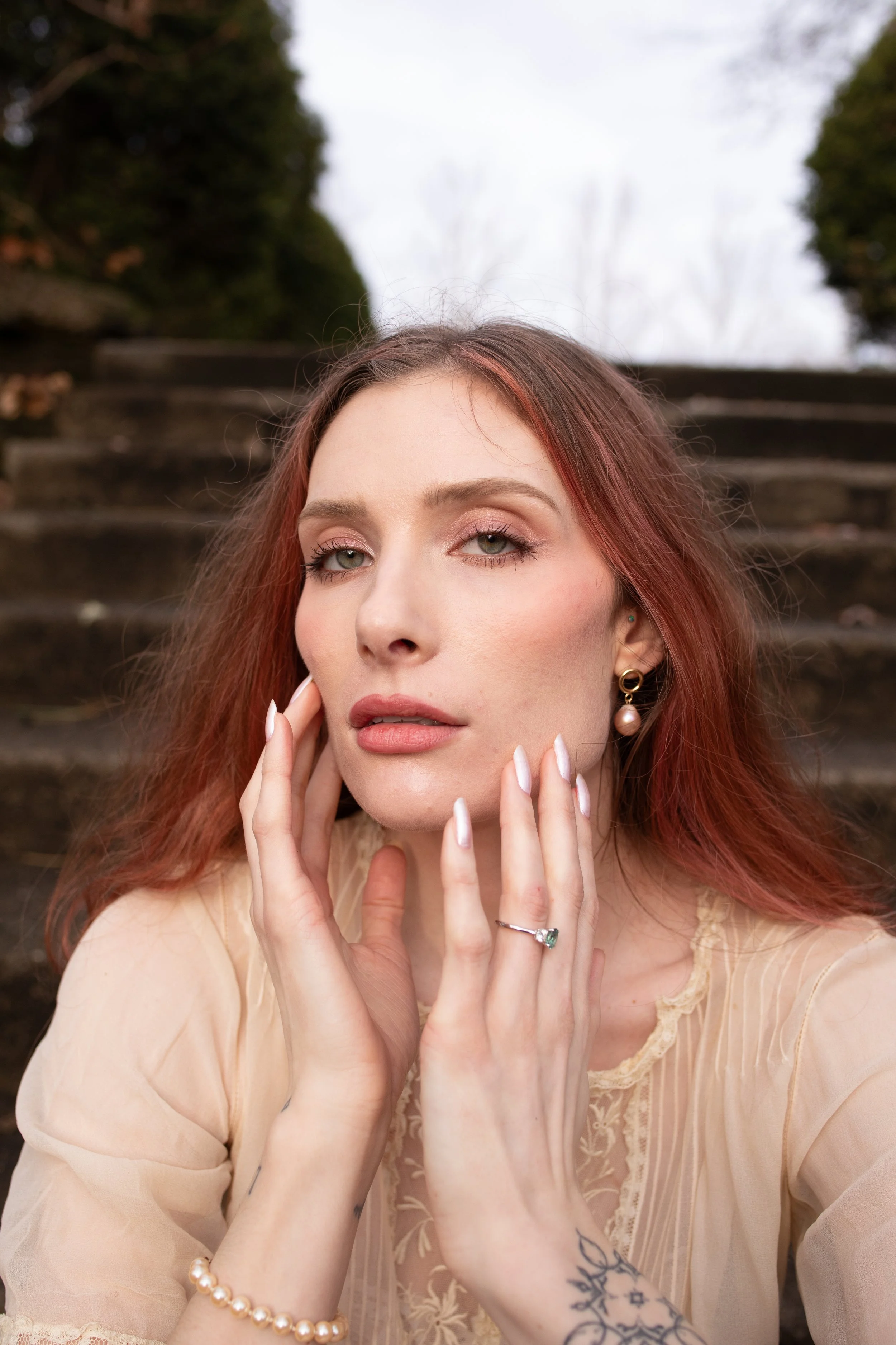 A close-up portrait of a woman with long red hair, green eyes, and makeup, outdoors on stone steps with trees and sky in the background. She is wearing a beige lace dress and jewelry including earrings, a ring, and a pearl bracelet.