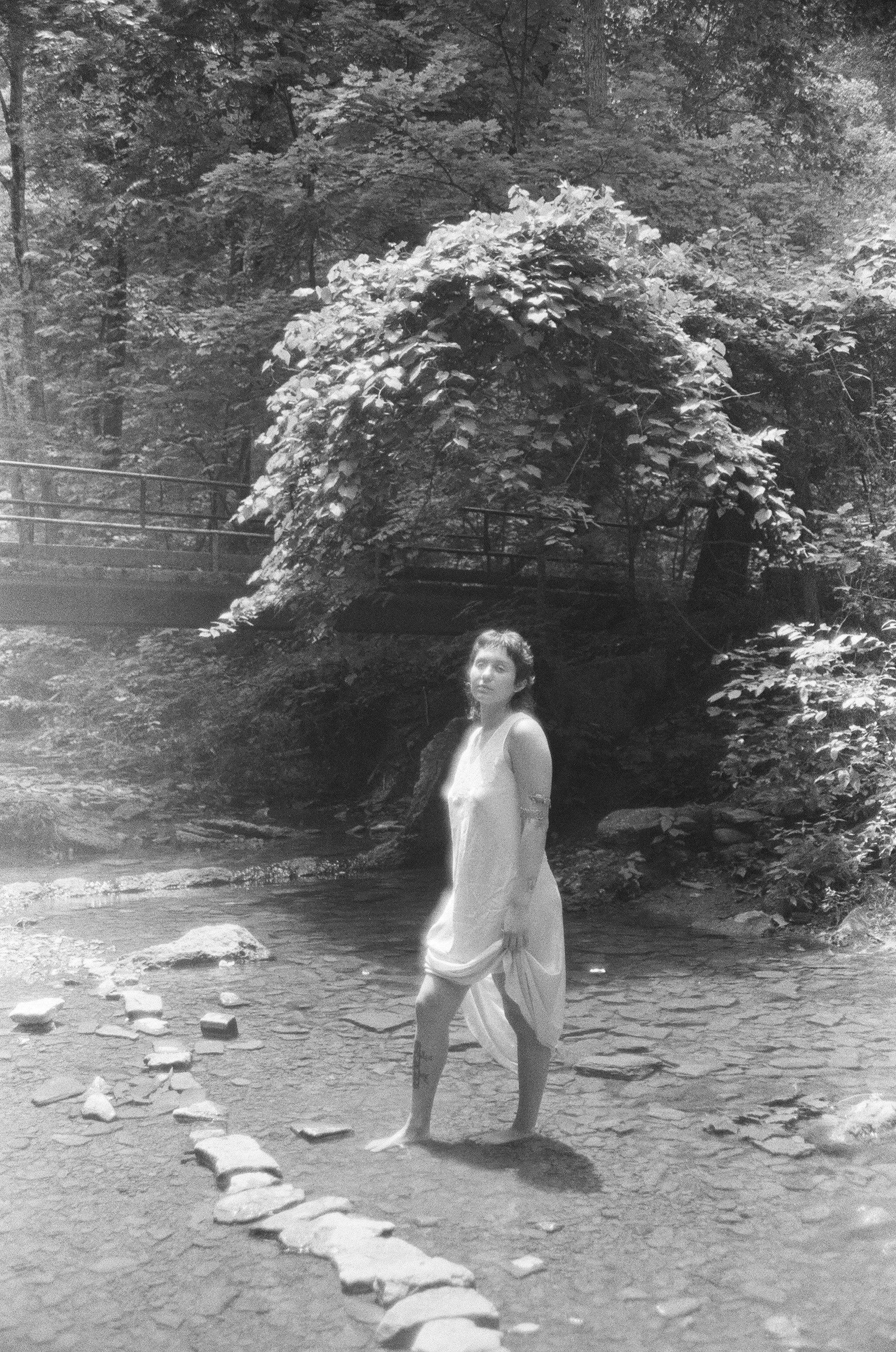 A young woman in a white dress standing barefoot in a shallow stream, with a line of rocks leading across the water, surrounded by trees and foliage.